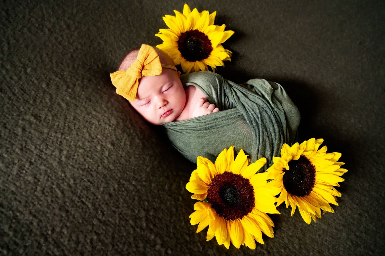 Sleeping newborn surrounded by bright yellow sunflowers on dark background.