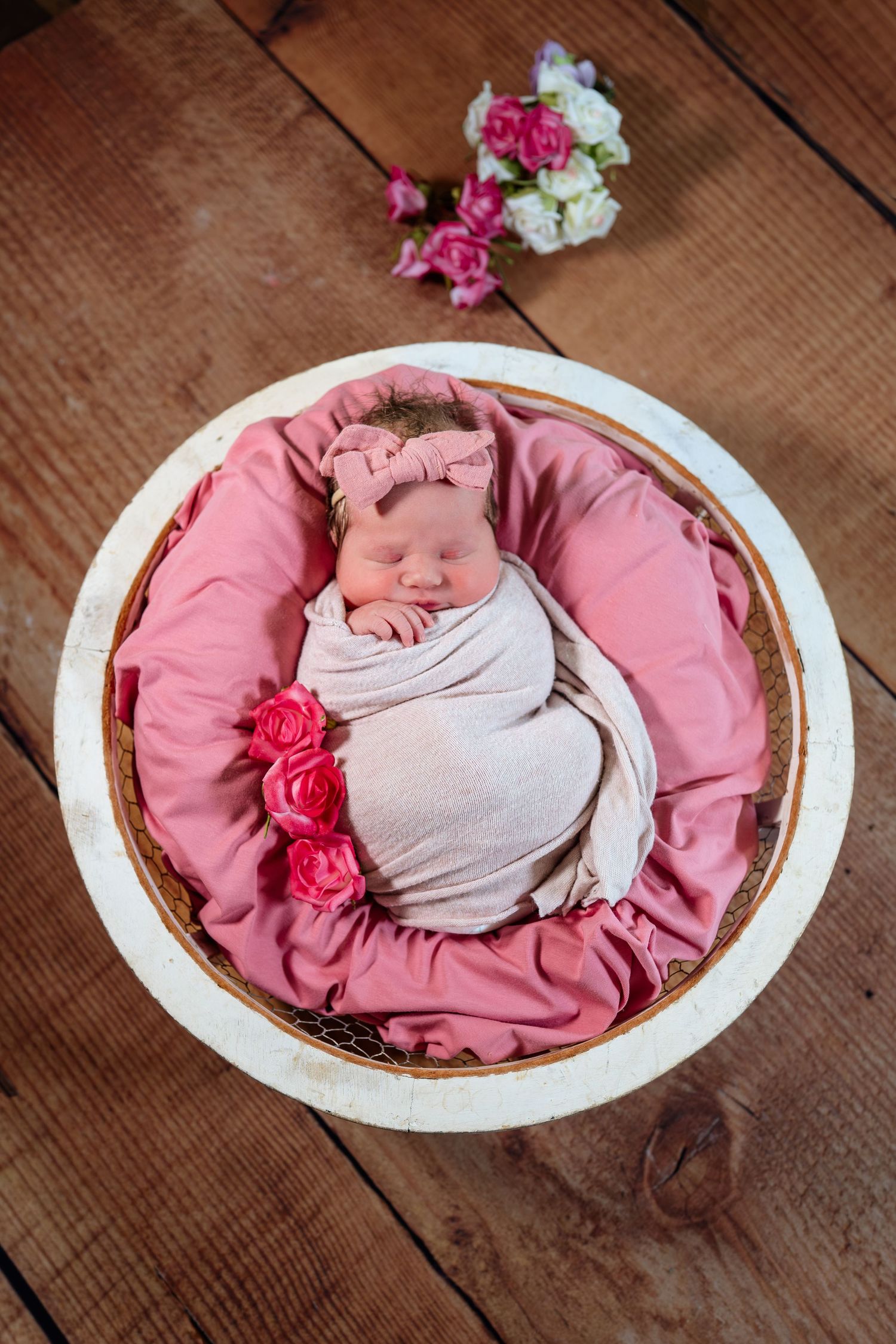 Newborn sleeping in pink oval basket decorated with roses on wooden surface.