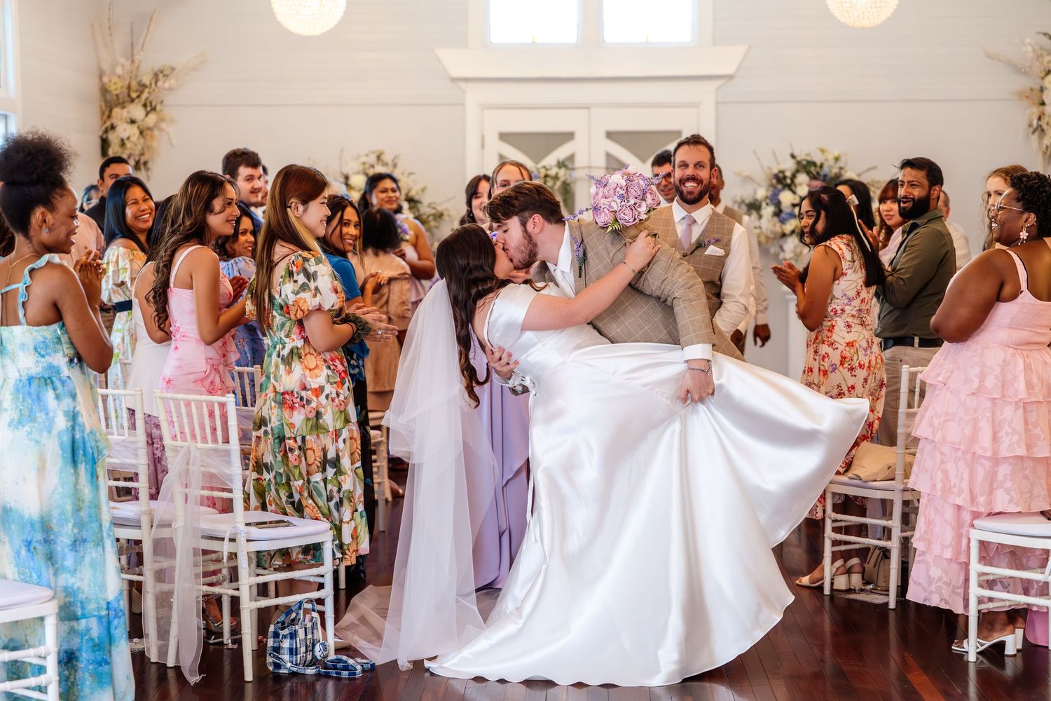 Bride and groom share a romantic first kiss at the altar surrounded by joyful wedding guests in a bright ceremony venue.