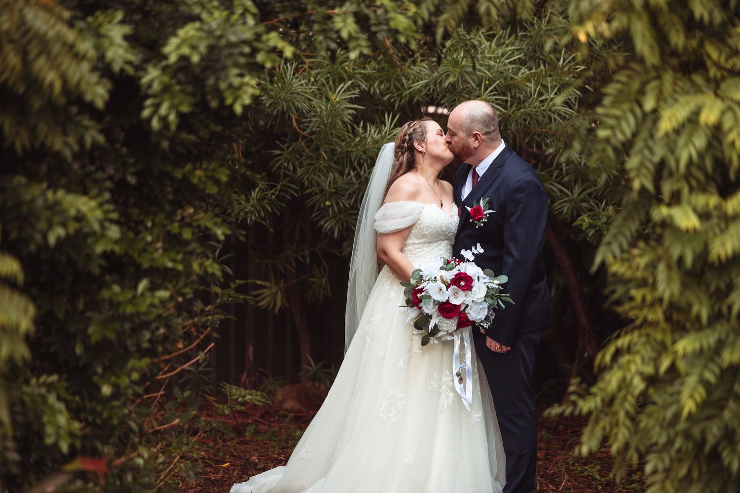 Bride and groom share a romantic kiss surrounded by lush green trees, bride holding a red and white bouquet.