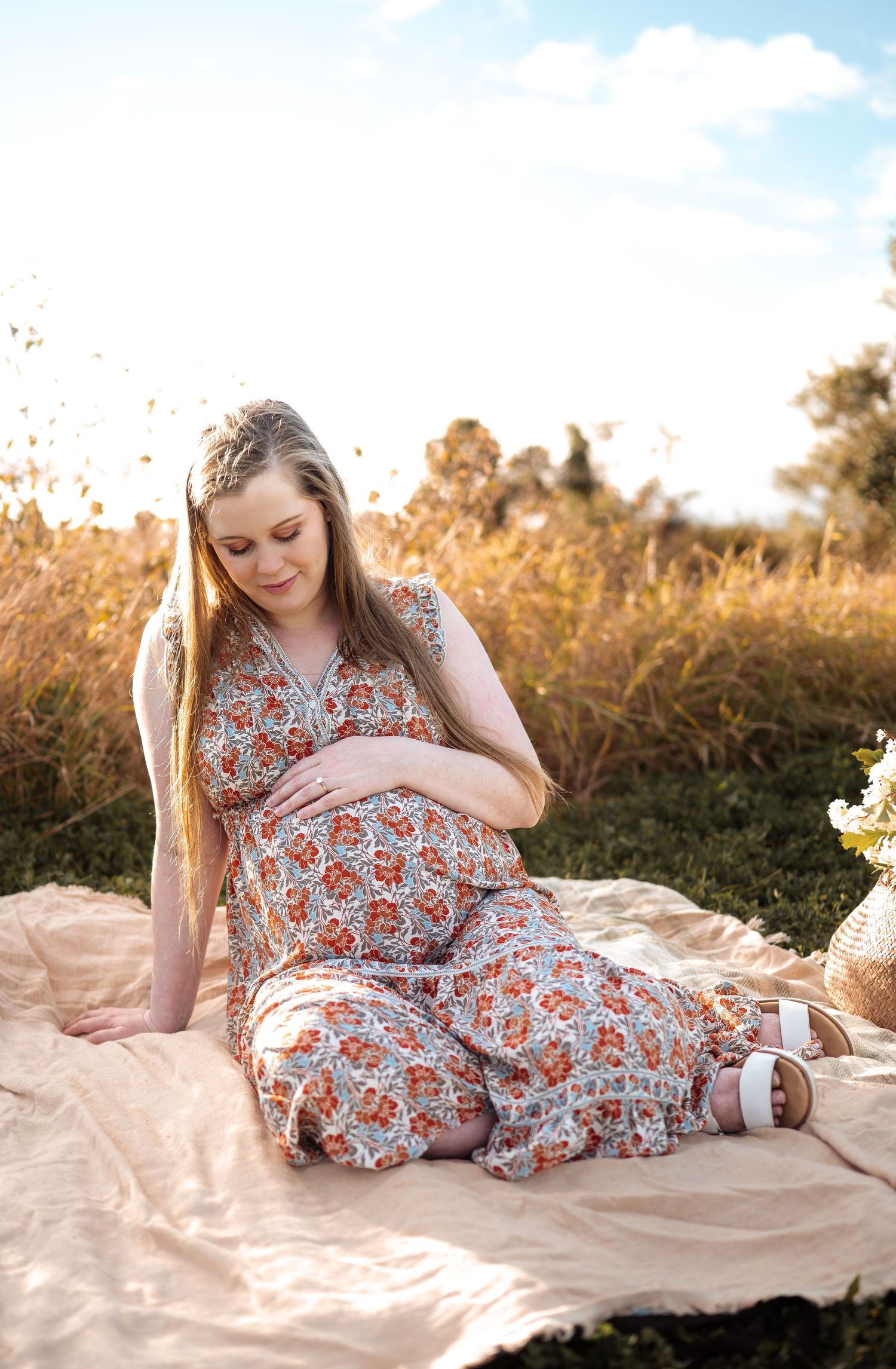 Expectant mother in floral dress reclining on blanket during golden hour outdoor photoshoot.