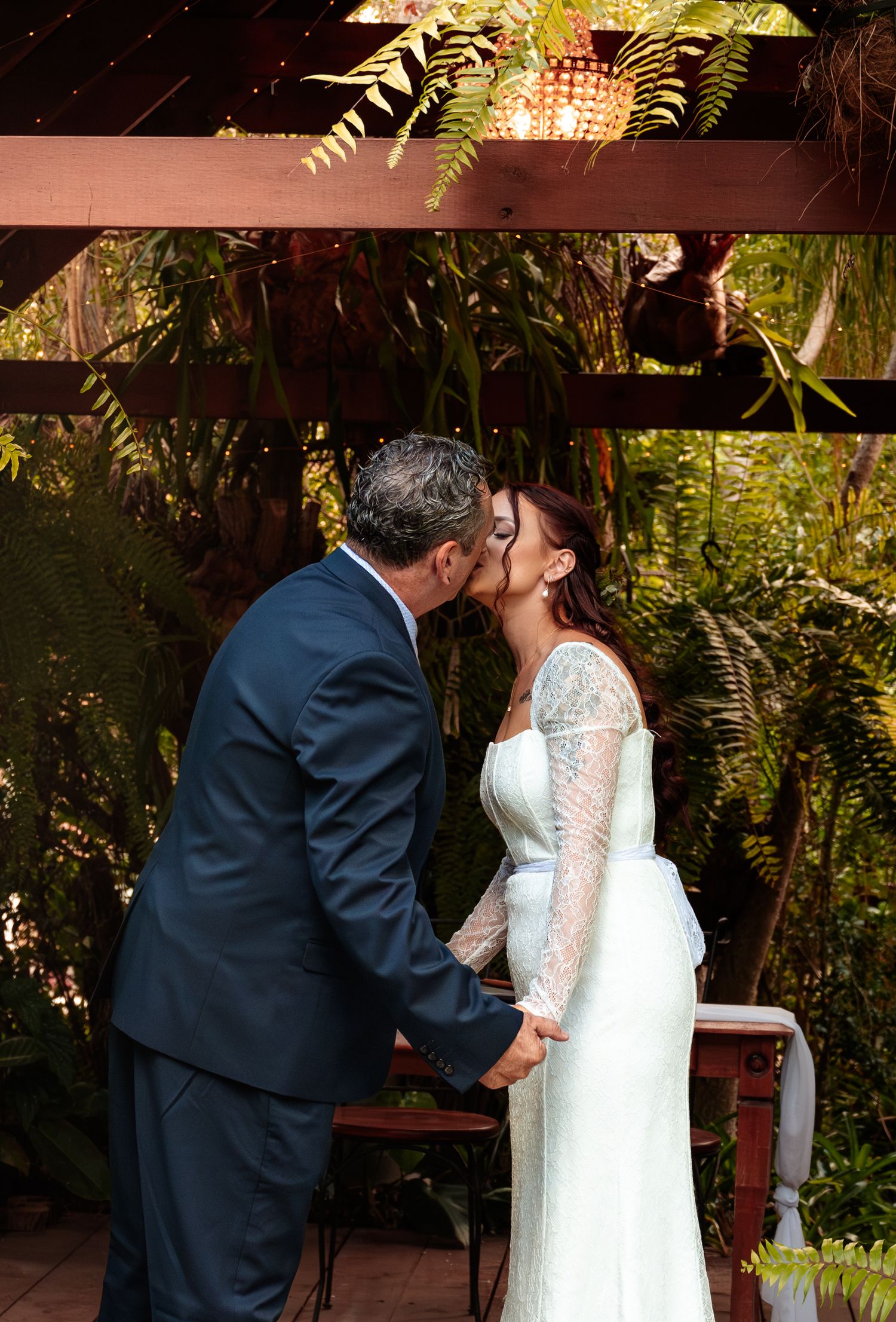 Bride and groom share their first kiss under a wooden pergola surrounded by lush tropical greenery.