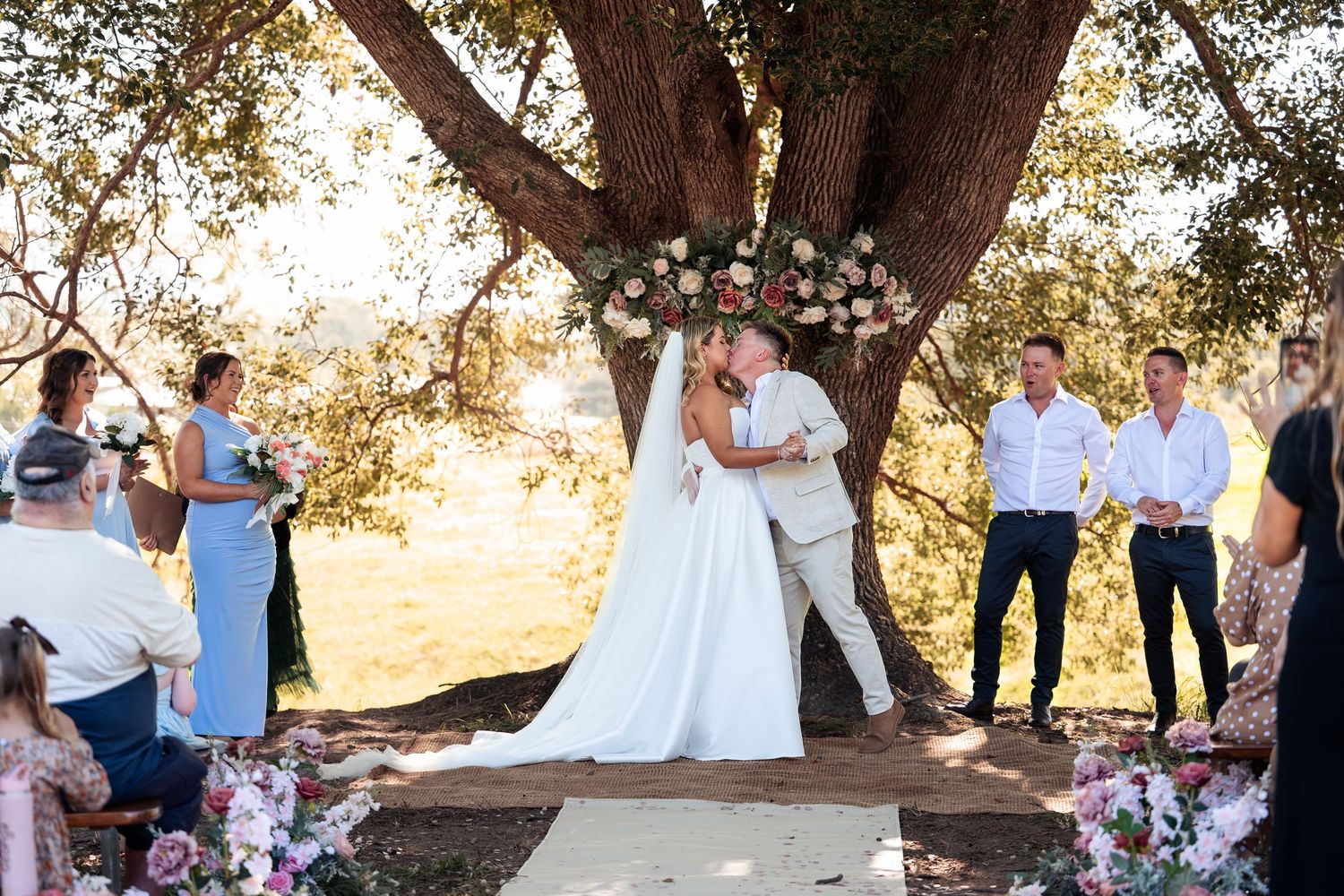 Couple shares first kiss at outdoor wedding ceremony under large oak tree with floral arch and guests gathered around.