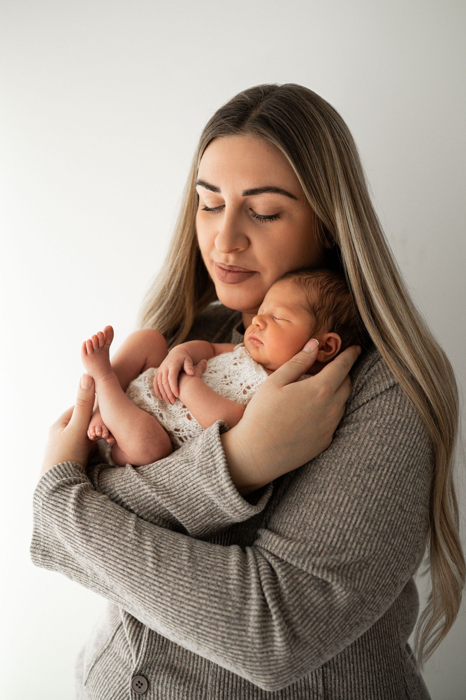 A blonde mother in a grey cardigan gently cradles her sleeping newborn baby against a white background.