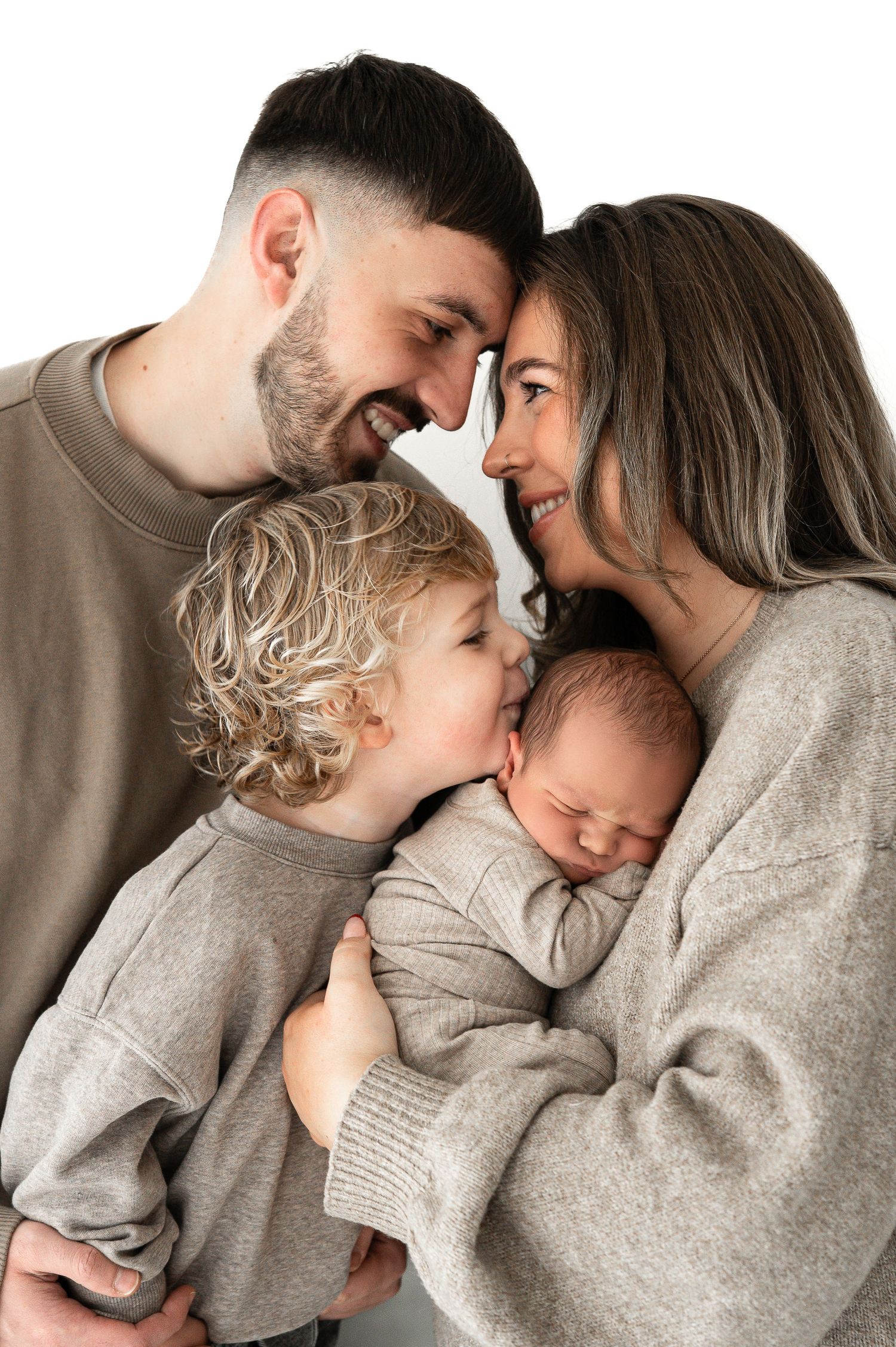 A happy family of four with parents smiling as their toddler kisses their sleeping newborn baby.