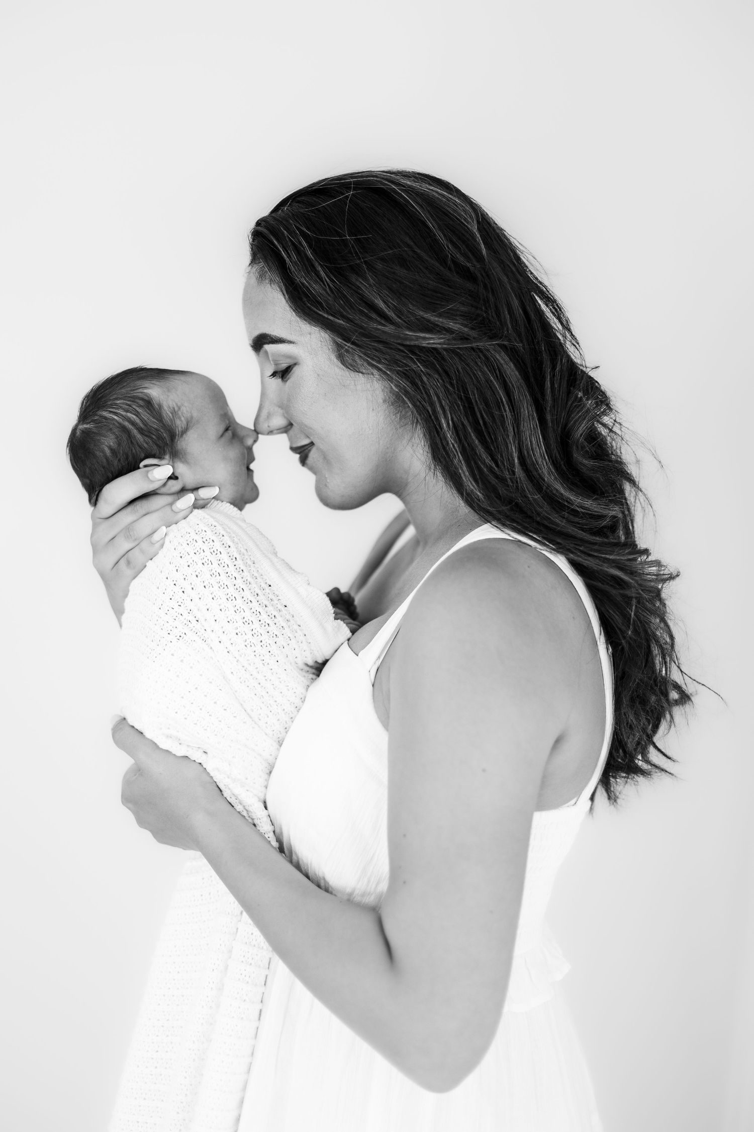 Black and white portrait of smiling mother in white dress tenderly holding swaddled newborn.
