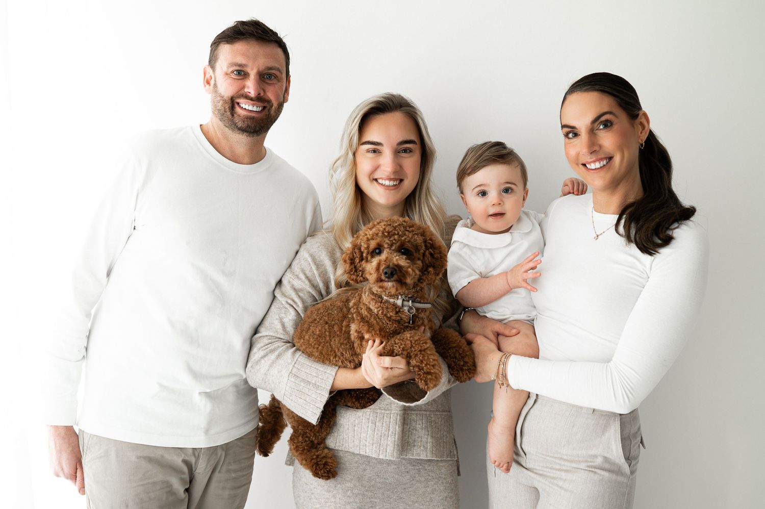 Happy family of four with curly brown dog and baby posing together in white outfits.