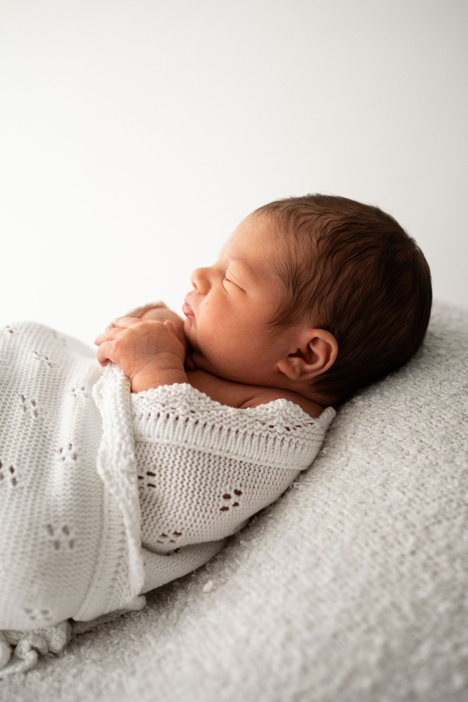 Close-up of a peaceful sleeping newborn wrapped in a white knit blanket against a soft grey surface.