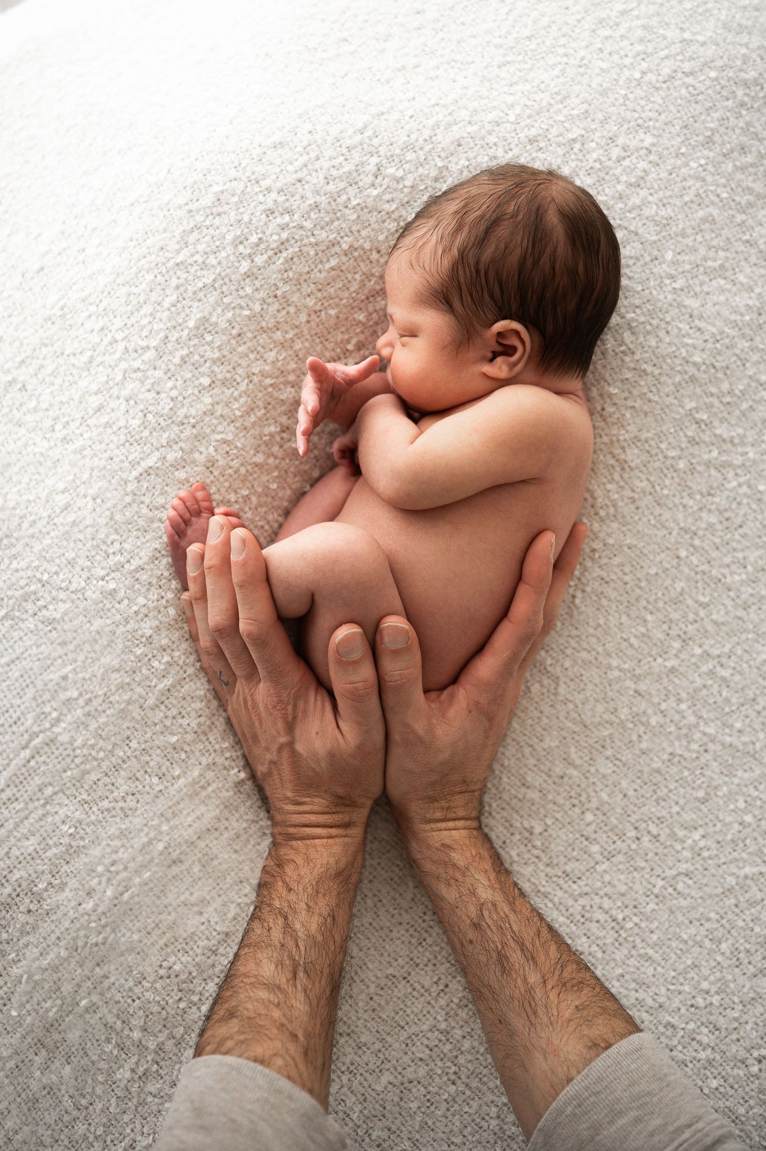 Father's hands gently cradle a sleeping naked newborn baby on a soft white blanket.