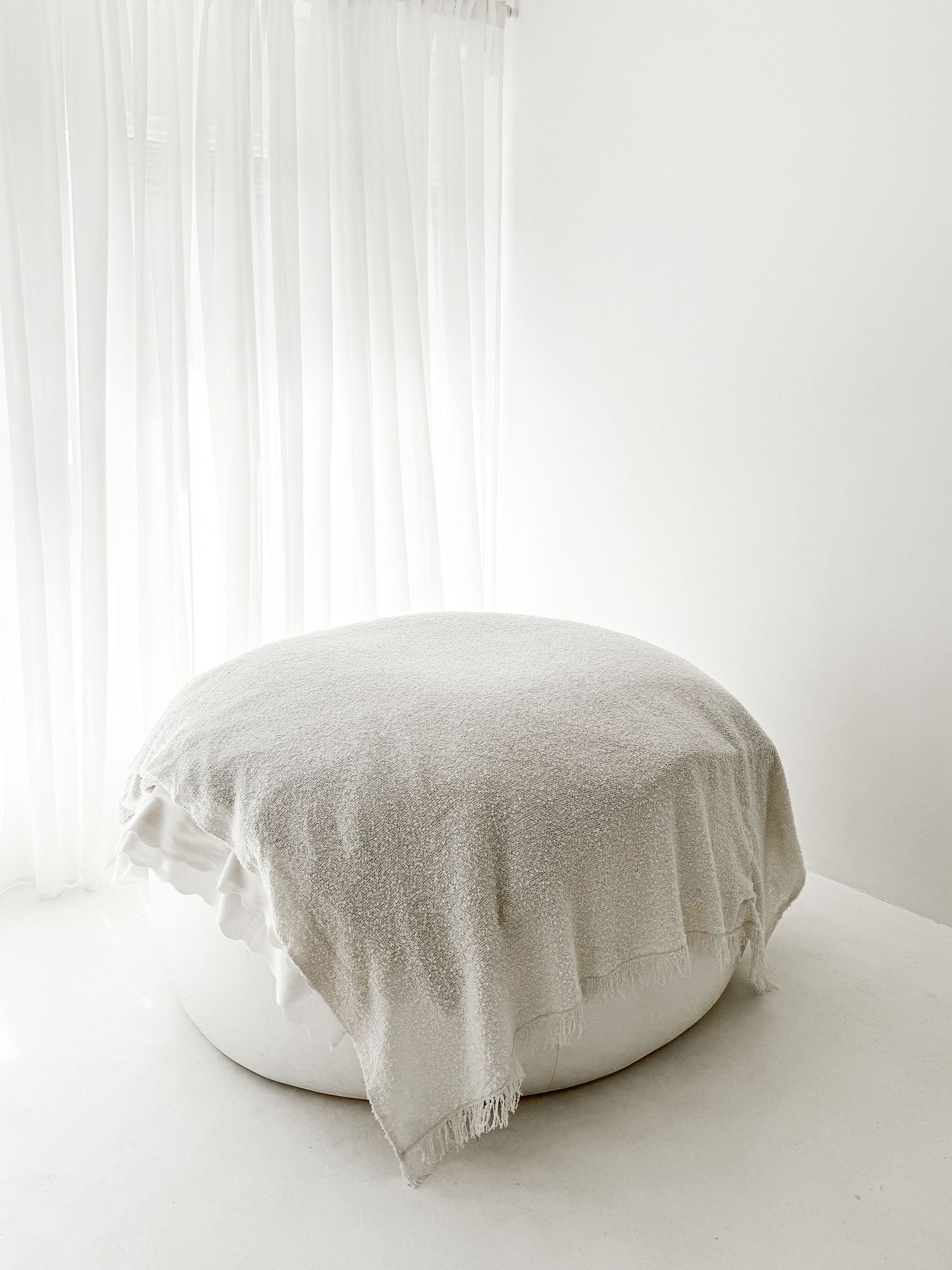 Round white pouf draped with fringed linen blanket in front of sheer white curtains.
