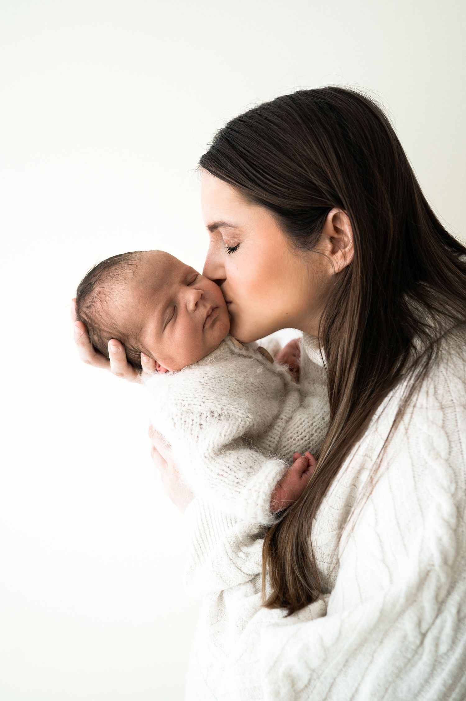 A dark-haired mother gently kisses her sleeping newborn baby wrapped in a white knit blanket.