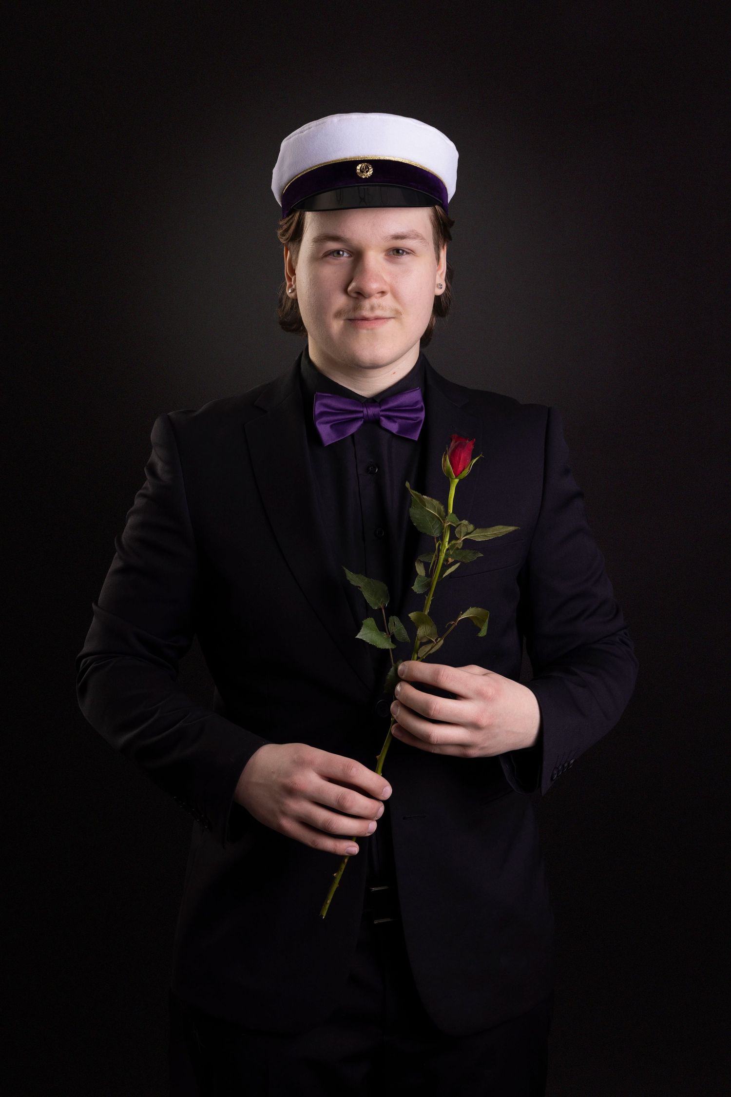 Mies poseeraa studiossa ylioppilas lakissa ja ruusu kädessä.