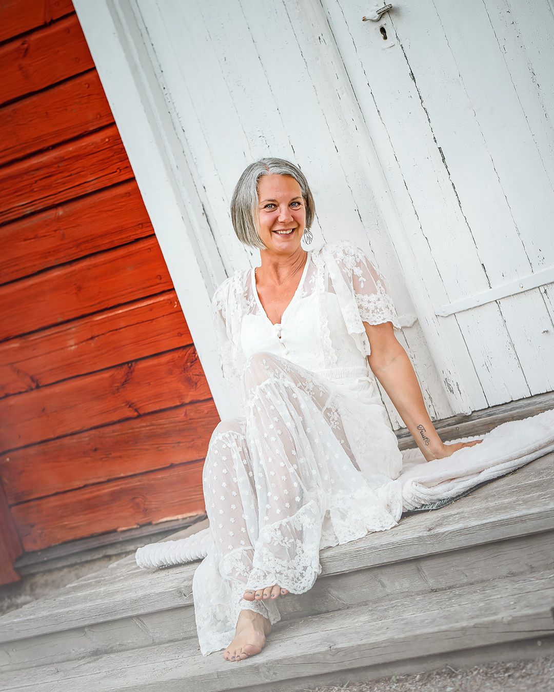 Smiling older woman with gray hair wearing a white lace dress, sitting barefoot on wooden steps near a red barn.