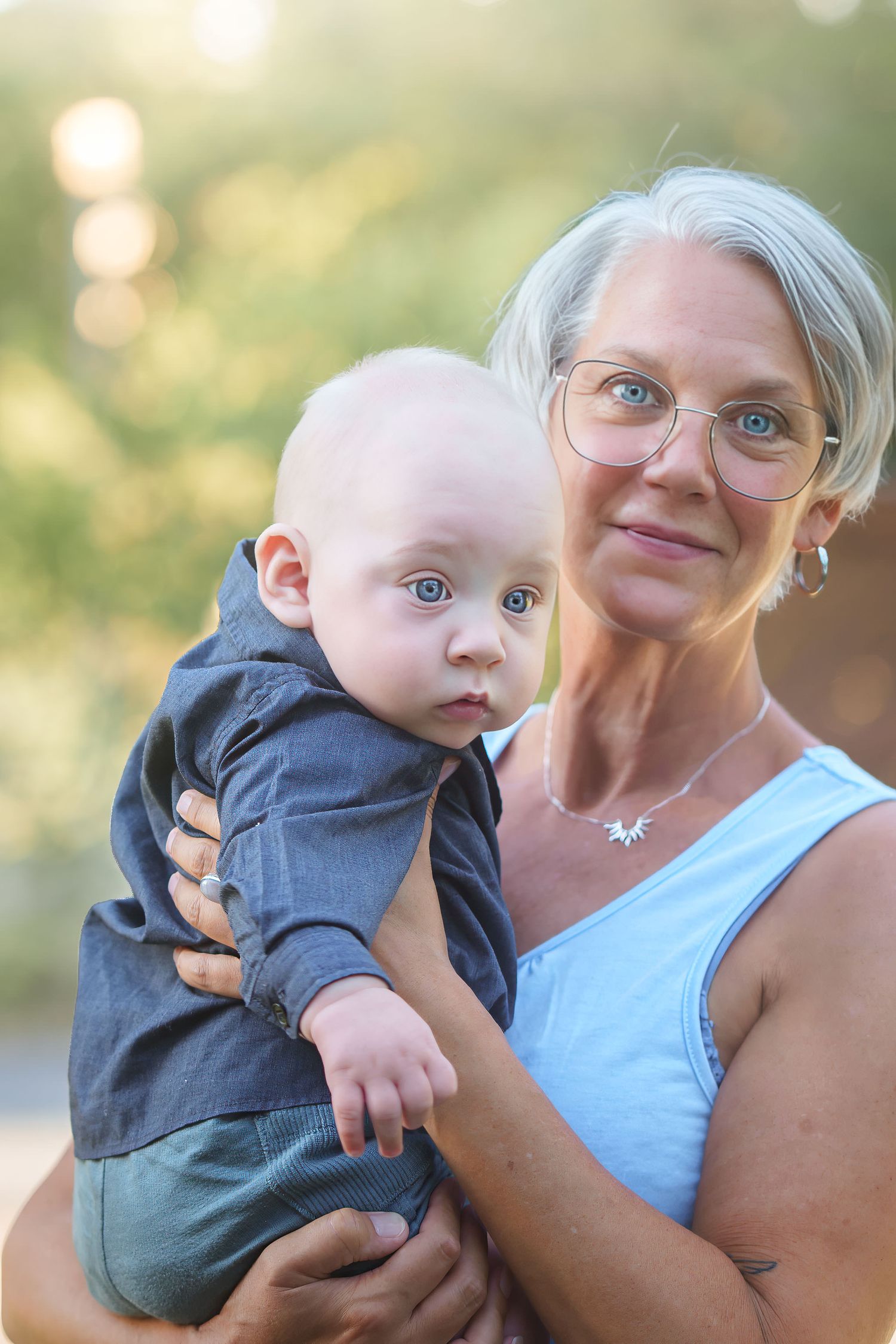 Grandmother with gray hair and glasses holds a baby boy outdoors in soft natural light.