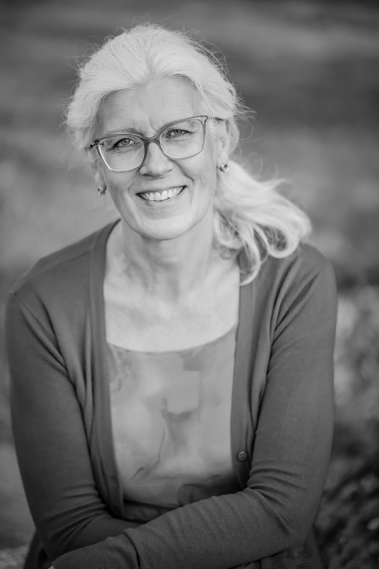 Smiling middle-aged woman with white hair and glasses posing outdoors in a black and white portrait photo.