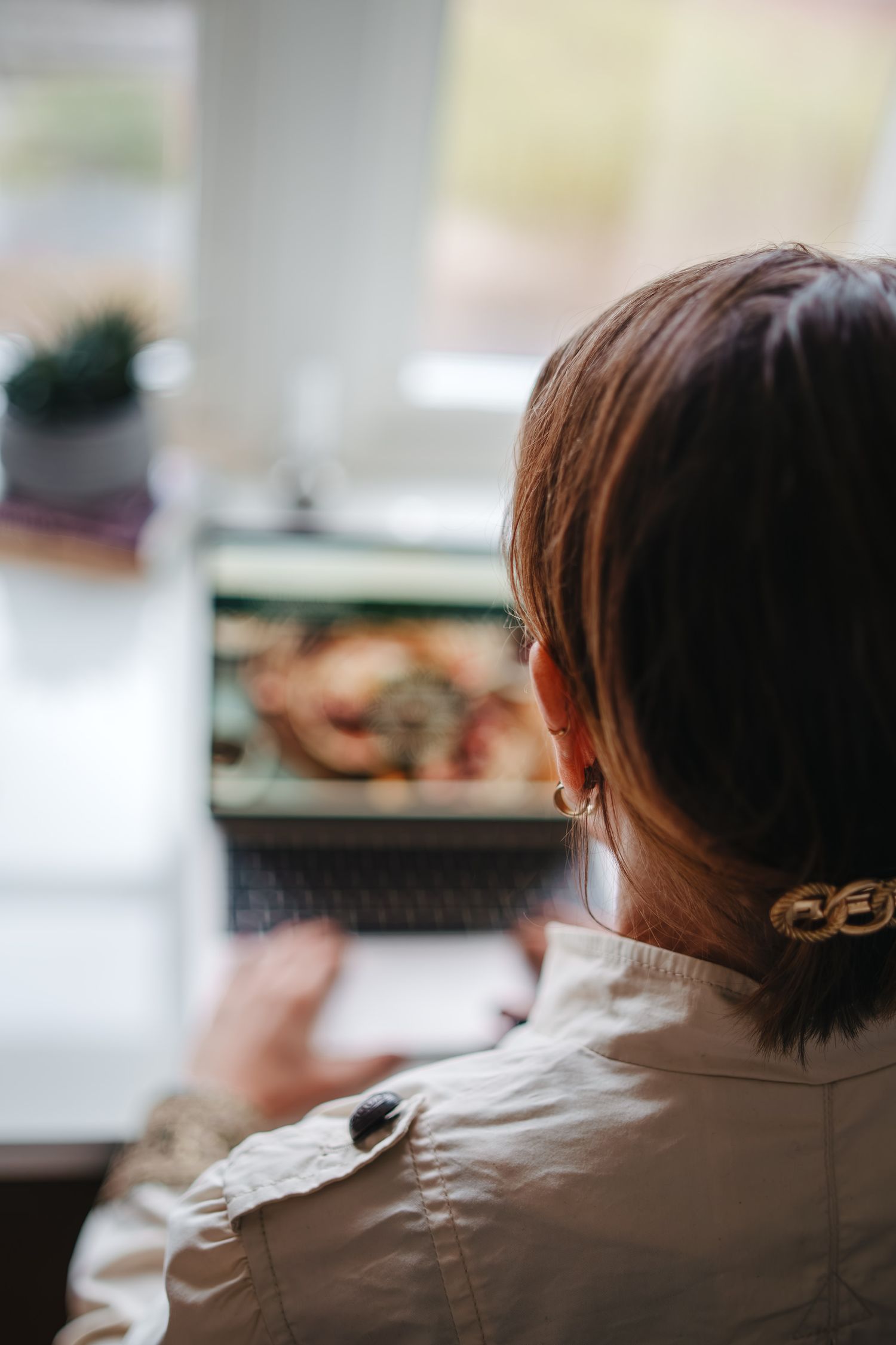 Woman with brown hair views food image on laptop near bright window, wearing beige jacket and gold hair clip.