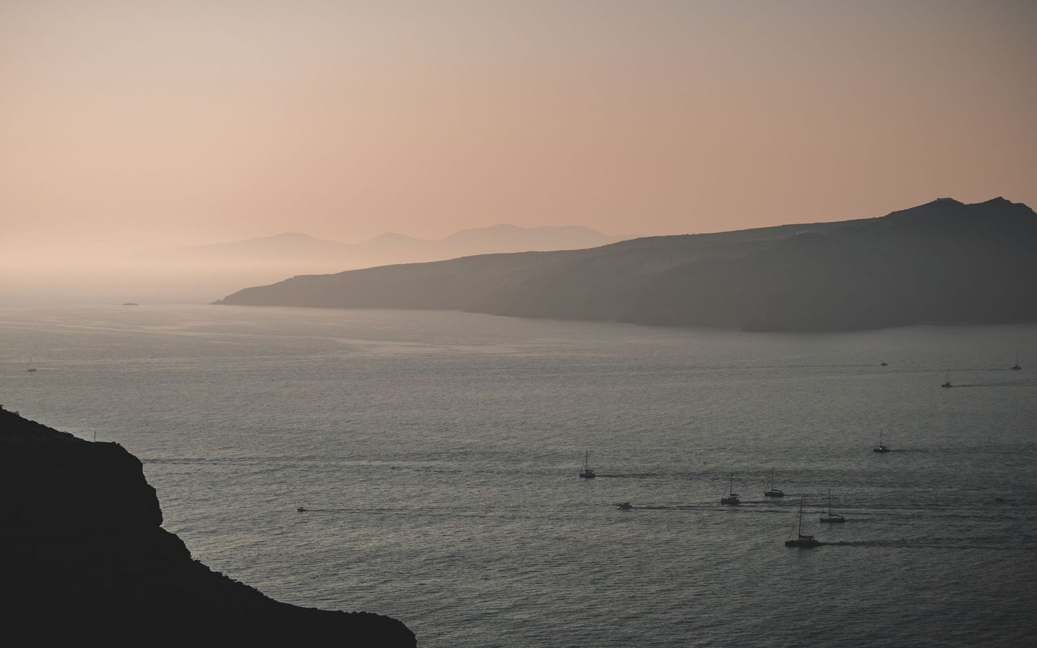Tranquil dusk seascape with silhouetted coastline and small boats dotting calm waters in Santorini.