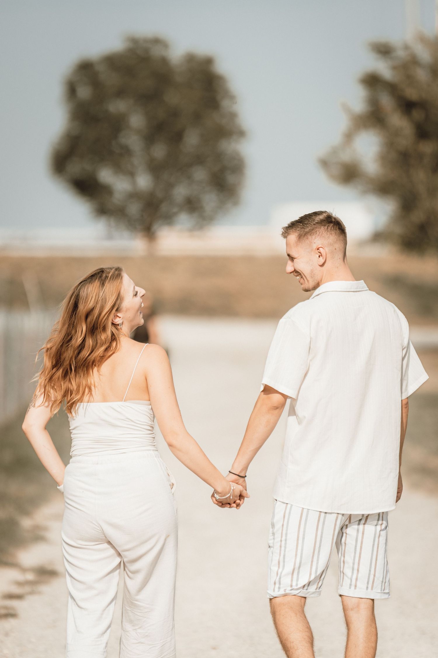 A smiling couple holding hands and walking on a sunny dirt path, dressed in white summer outfits.