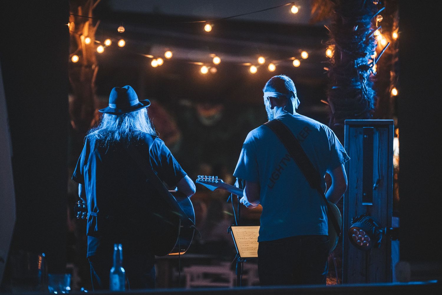 Two musicians performing on stage under warm string lights in a dark outdoor night venue.