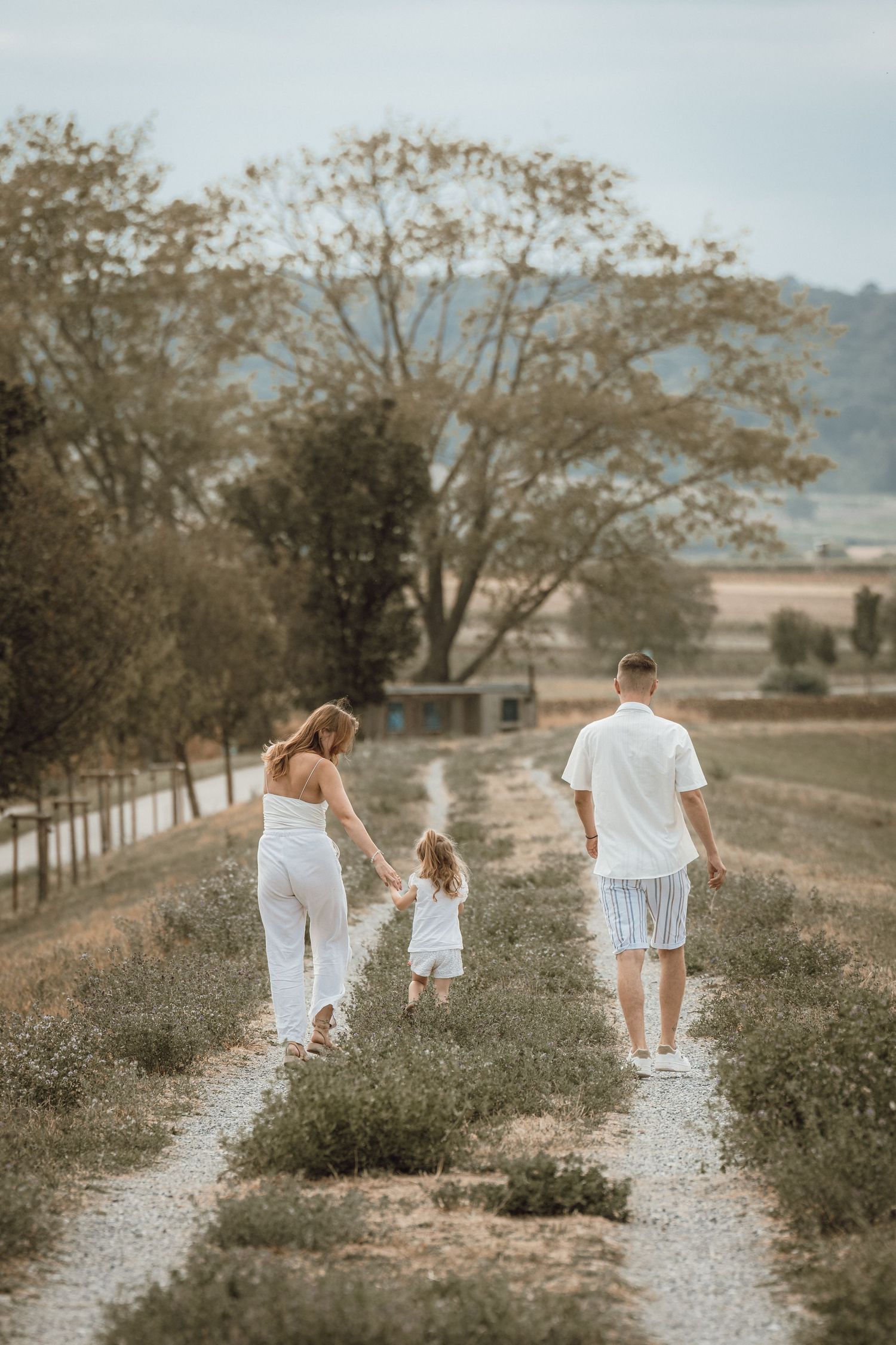 Family of three in white outfits walking hand-in-hand along a scenic countryside path surrounded by greenery.