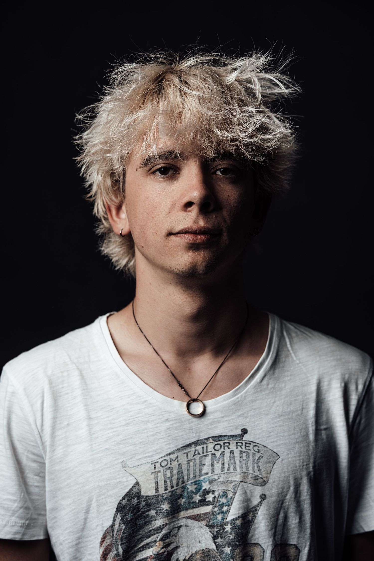 Young man with messy blonde hair wearing a white Trademark graphic tee and necklace poses against dark background.