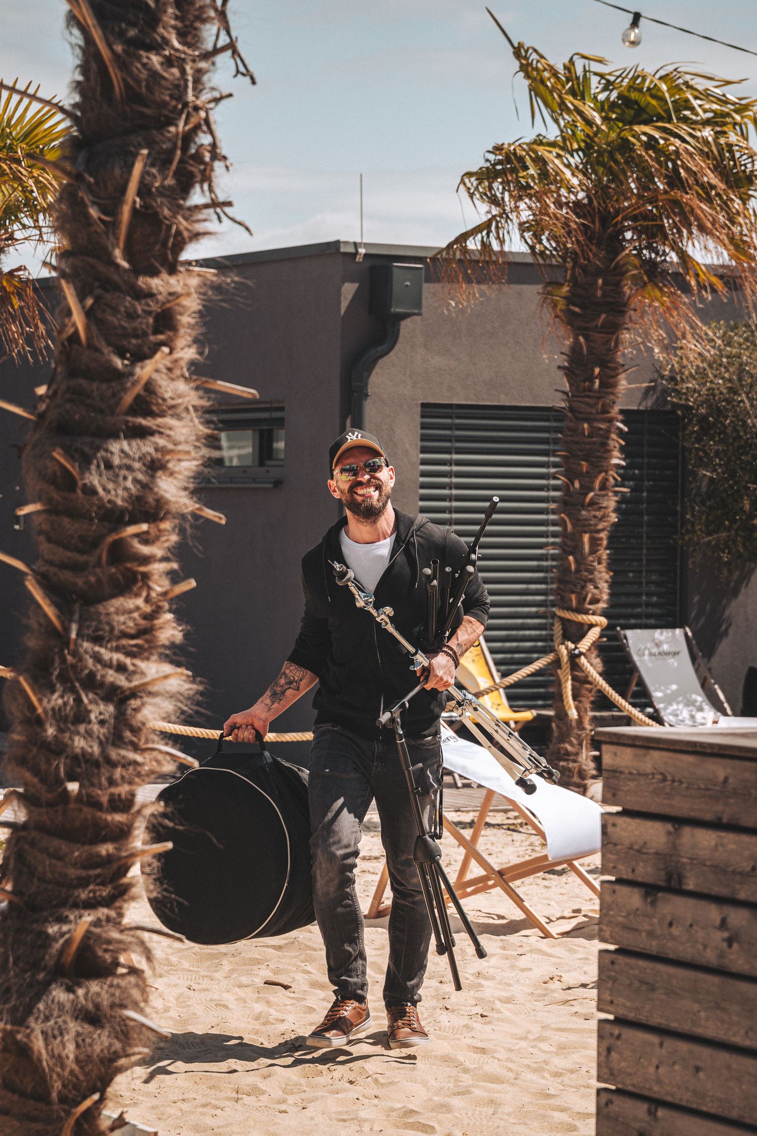 Smiling tattooed man carrying drum equipment between palm trees at a beach venue.
