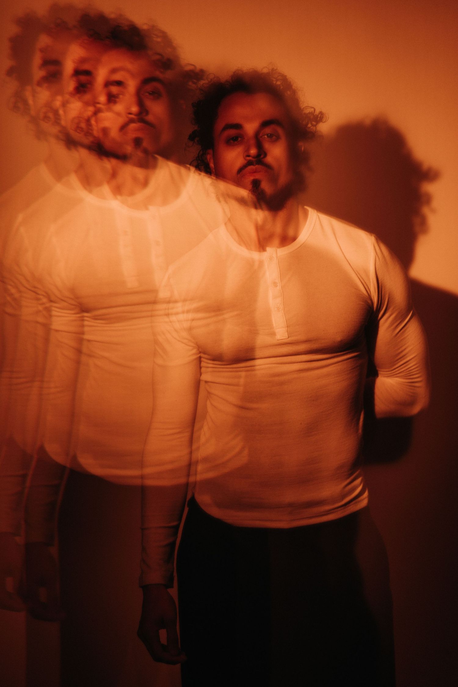 Multiple exposure portrait of a curly-haired bearded man in a white henley shirt, bathed in warm orange light with a dramatic shadow.