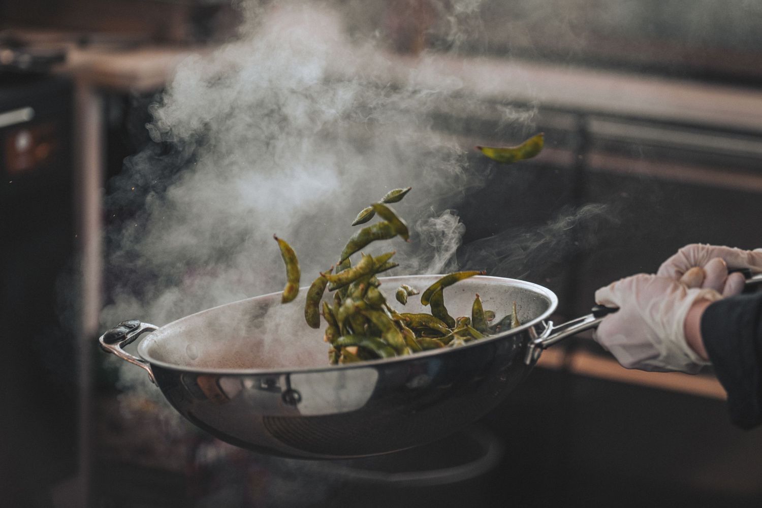 Chef tossing green vegetables in a steaming wok on a professional kitchen stove.