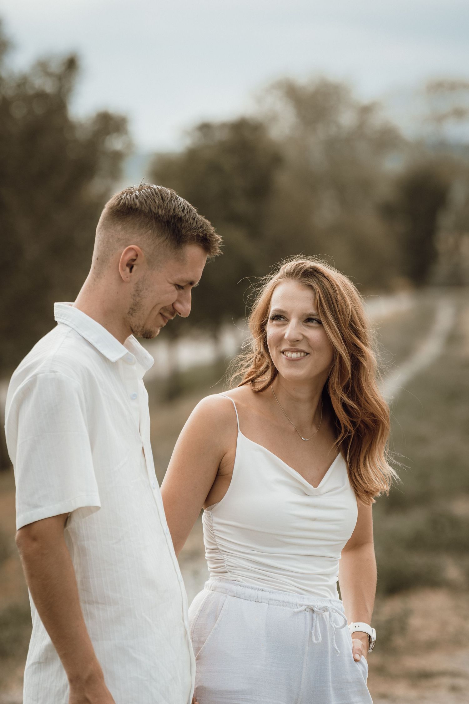 A smiling couple in matching white outfits standing outdoors in a natural setting with trees in the background.