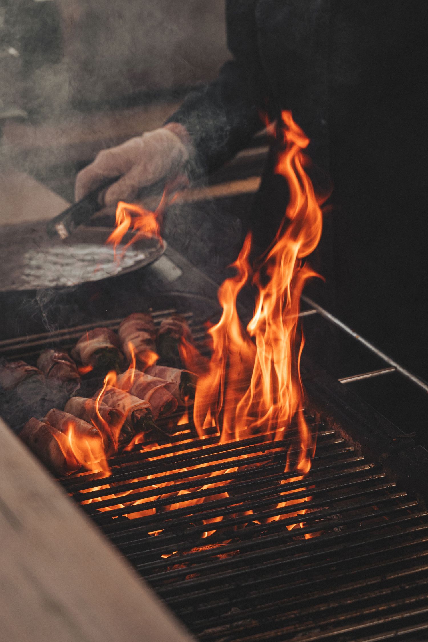 Flames rising from a charcoal BBQ grill with meat cooking and smoke filling the air.