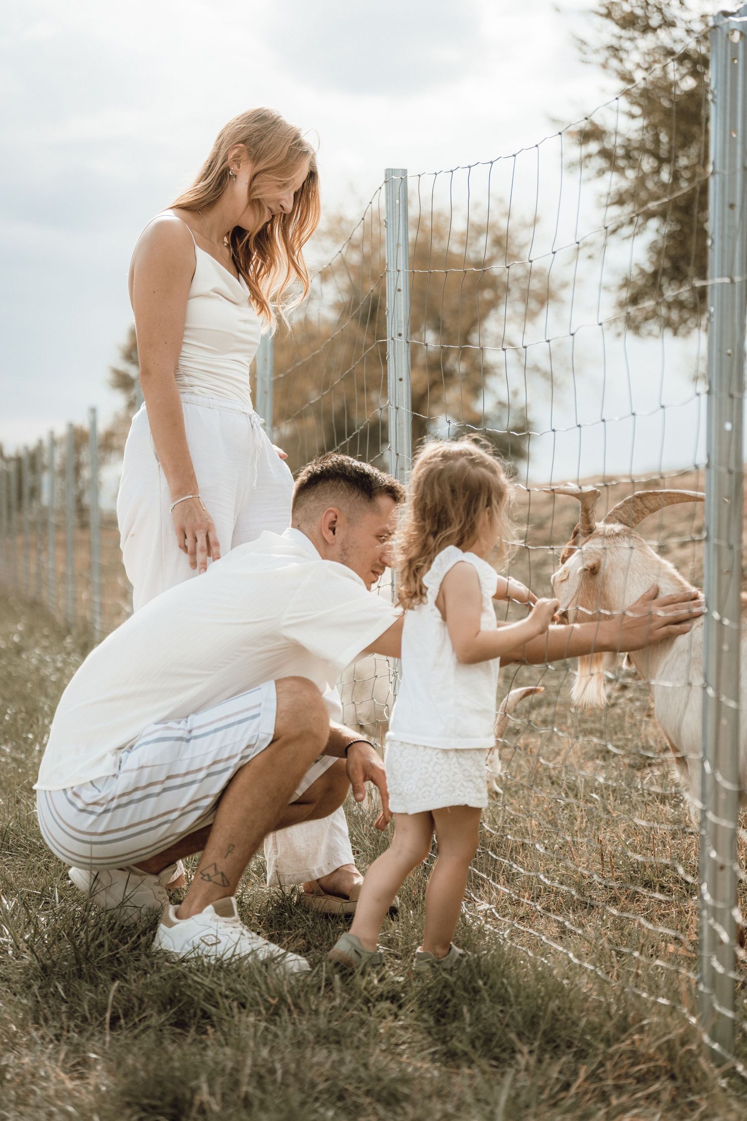 Family in white outfits feeding a goat through a wire fence on a sunny farm day.