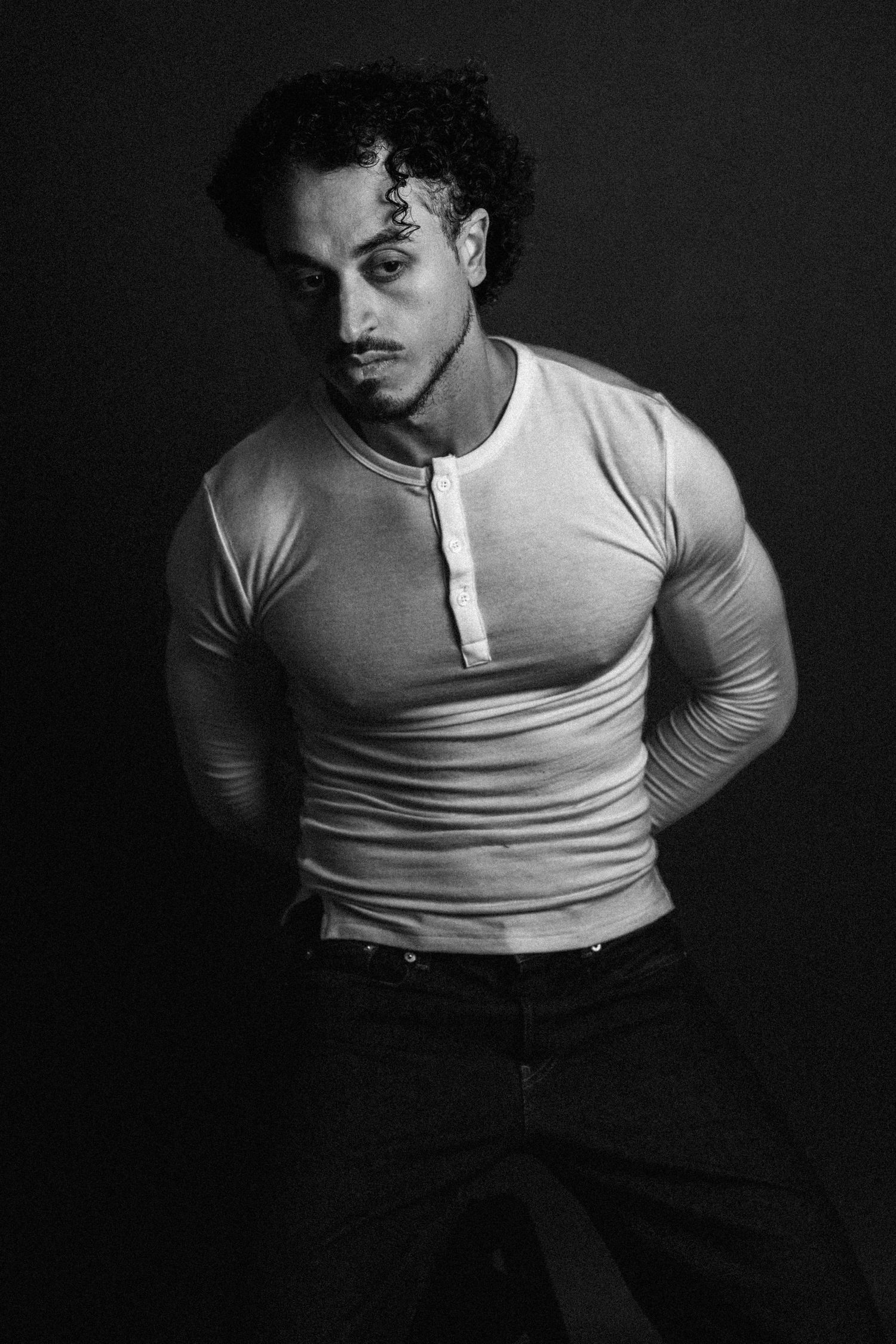 Black and white portrait of a curly-haired man with a goatee in a fitted henley shirt, posing with his hands behind his back.