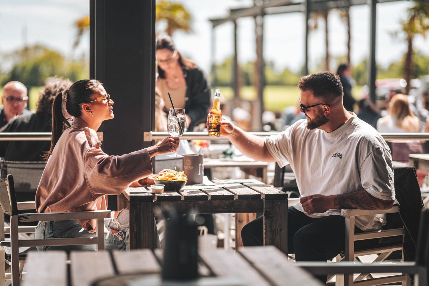 Man and woman toasting with drinks at a sunny outdoor restaurant terrace.