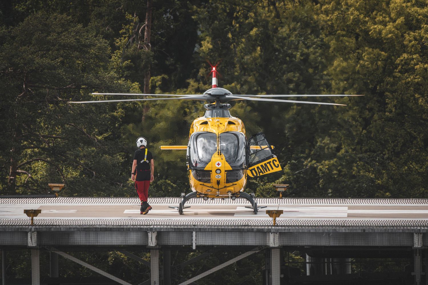 Yellow rescue helicopter on helipad with crew member standing nearby, surrounded by lush green trees.