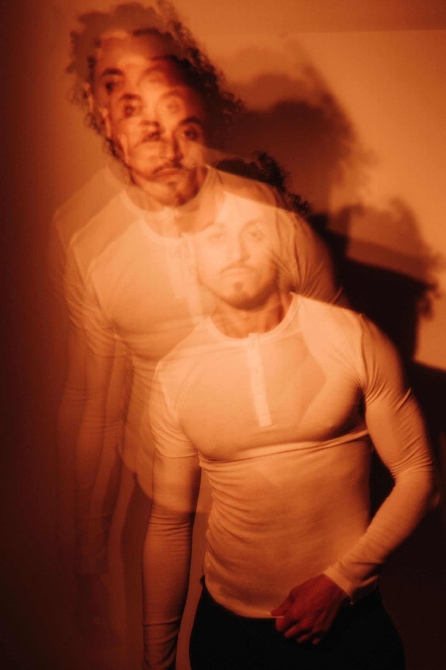 Multiple exposure portrait of a bearded man in a fitted white henley shirt, lit by warm orange light with ghostly overlapping images.