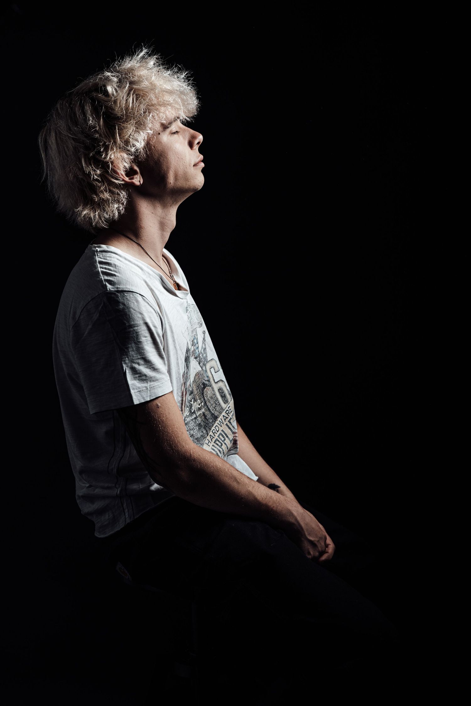 Young man with blonde hair tilts head back in dramatic low-key studio portrait against black background.