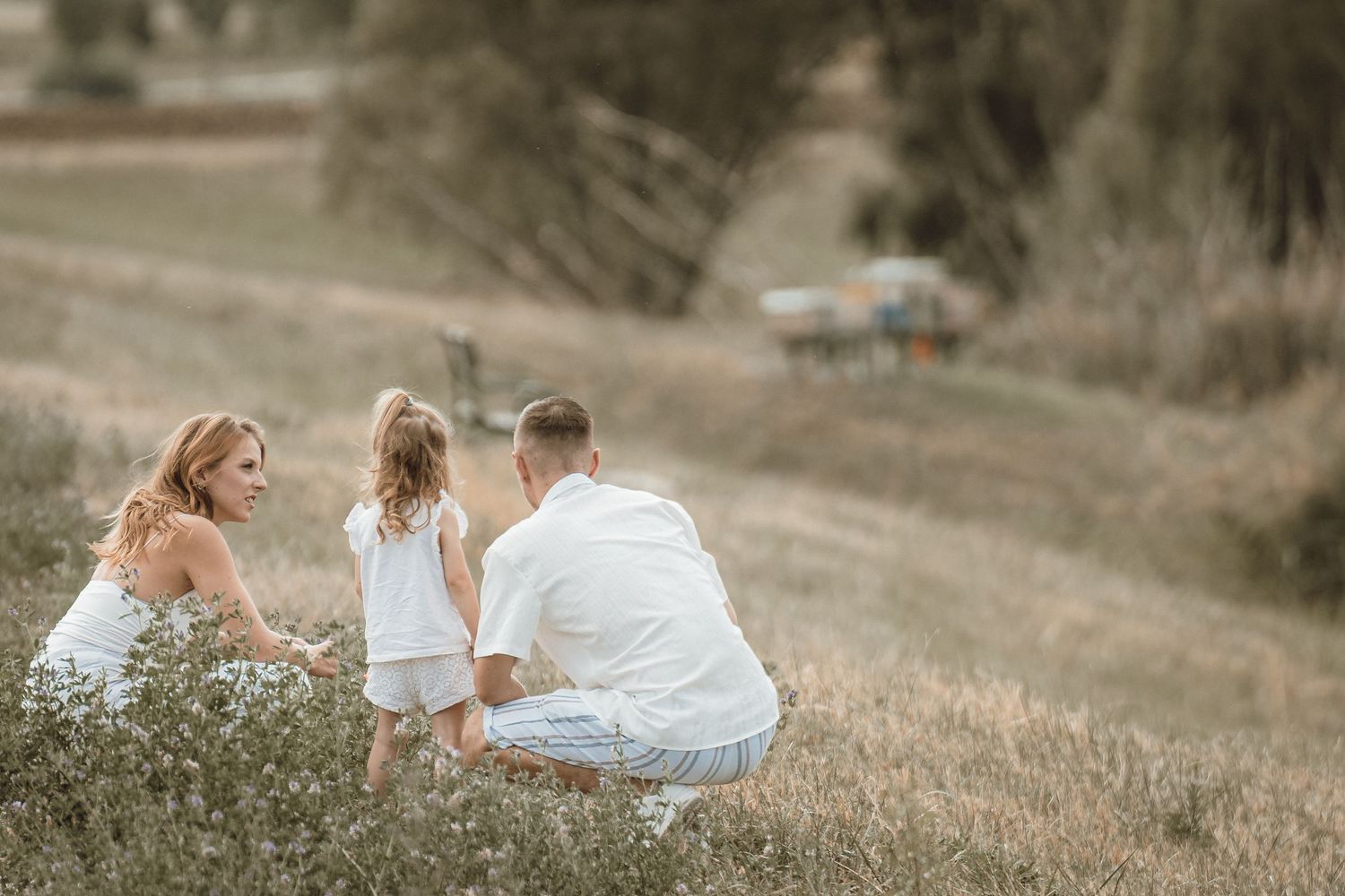 A family of three in white outfits enjoying a peaceful moment together in a sunlit meadow field.