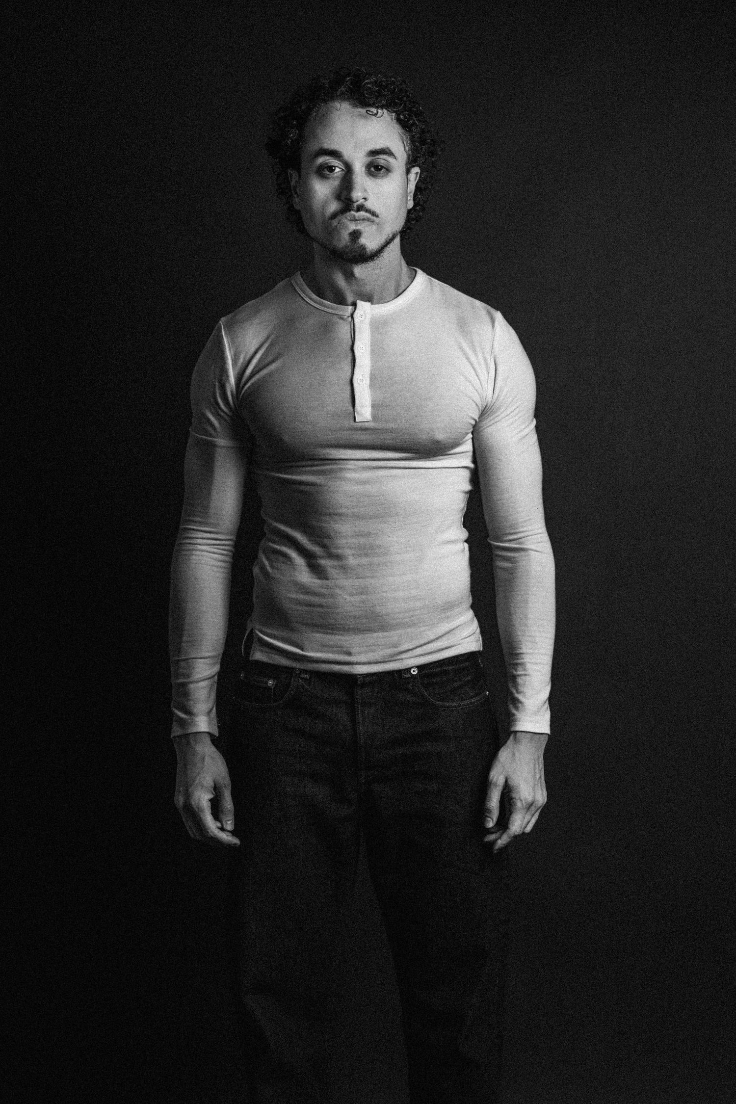 Black and white studio portrait of a curly-haired man with a goatee wearing a fitted henley shirt and dark jeans against a dark backdrop.