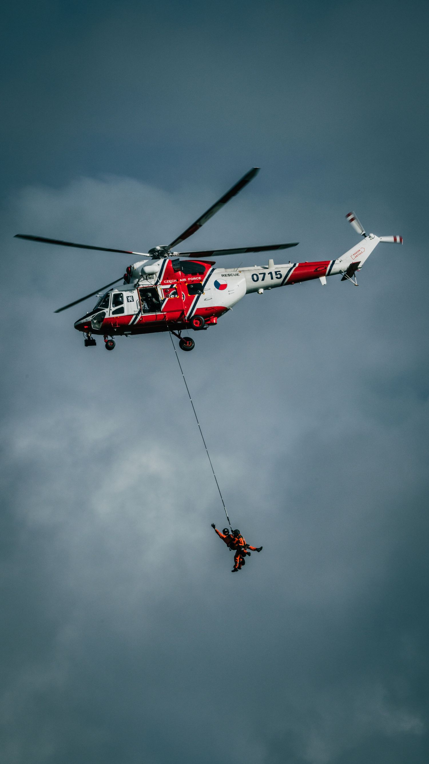 Red and white rescue helicopter number 0715 performing hoist rescue operation against cloudy sky.
