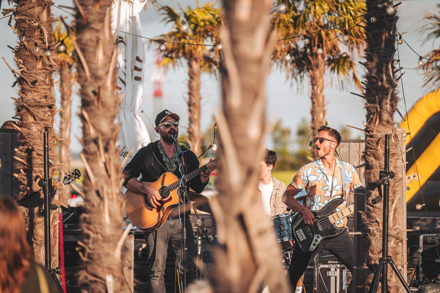 Two musicians performing live outdoors with acoustic and electric guitars among palm trees.