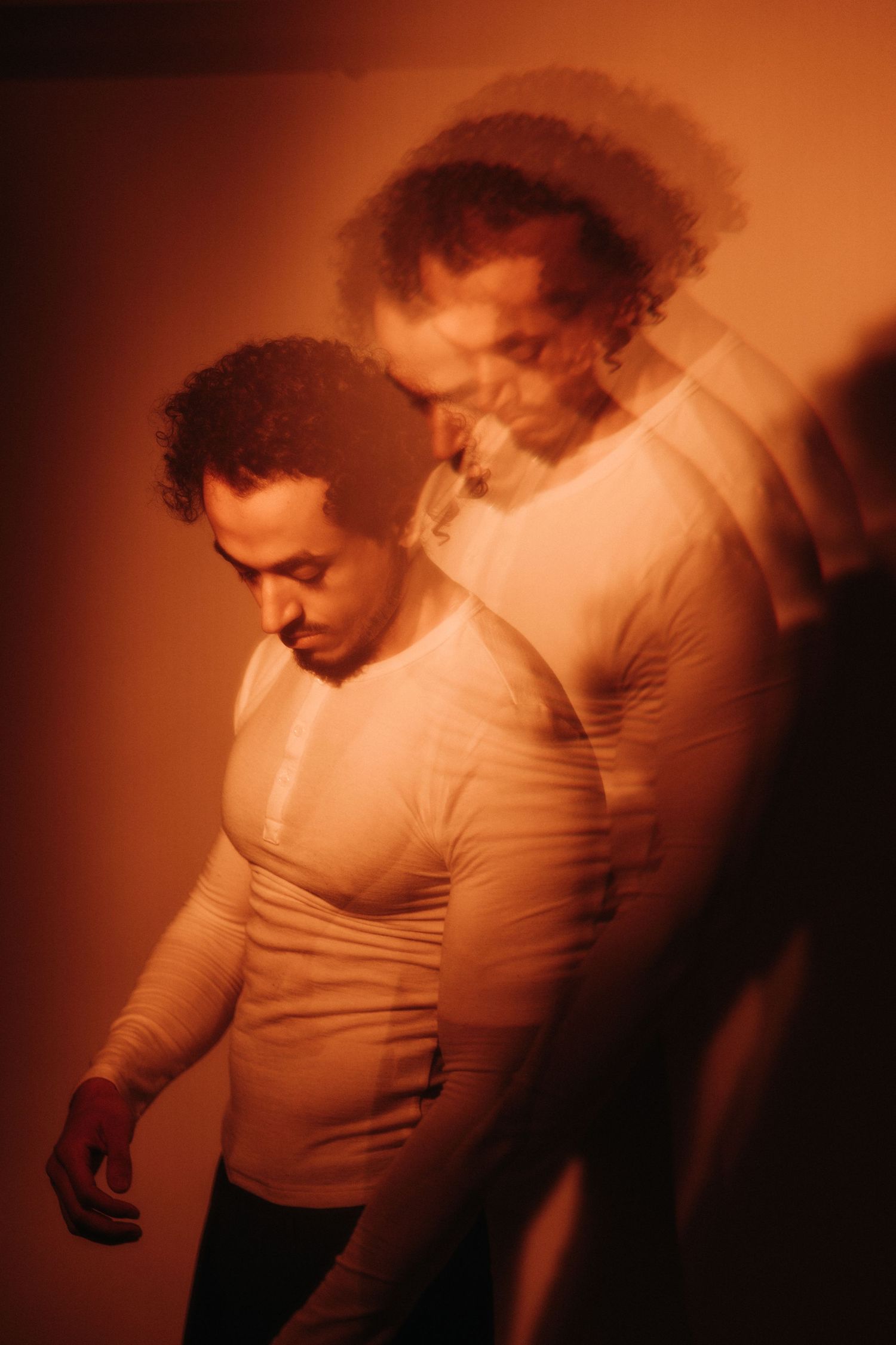 Multiple exposure portrait of a man with curly hair looking downward, wearing a fitted light shirt, bathed in warm orange light.