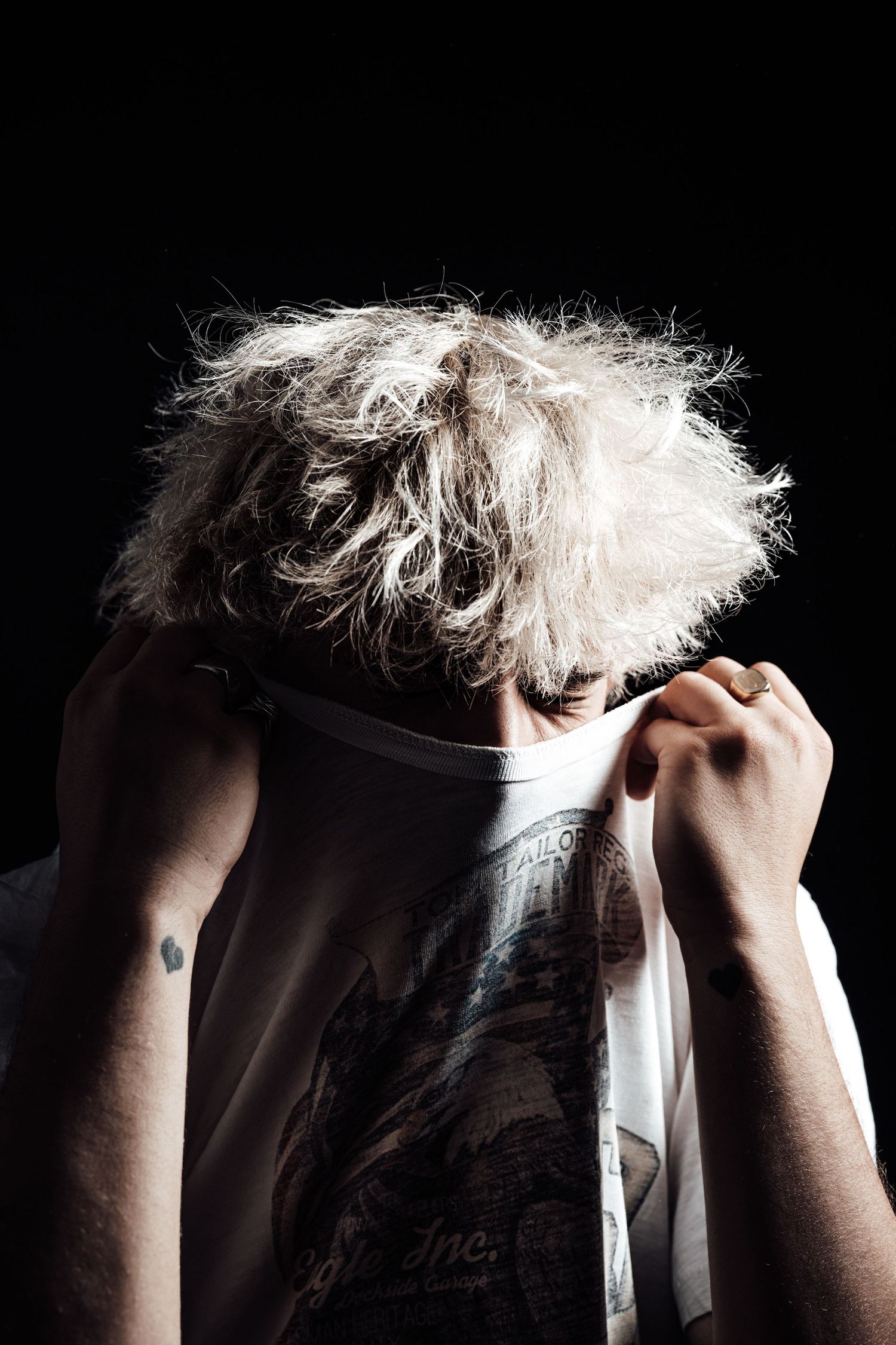 Person with wild, messy blonde hair pulling a white graphic t-shirt over their face against a dark background.
