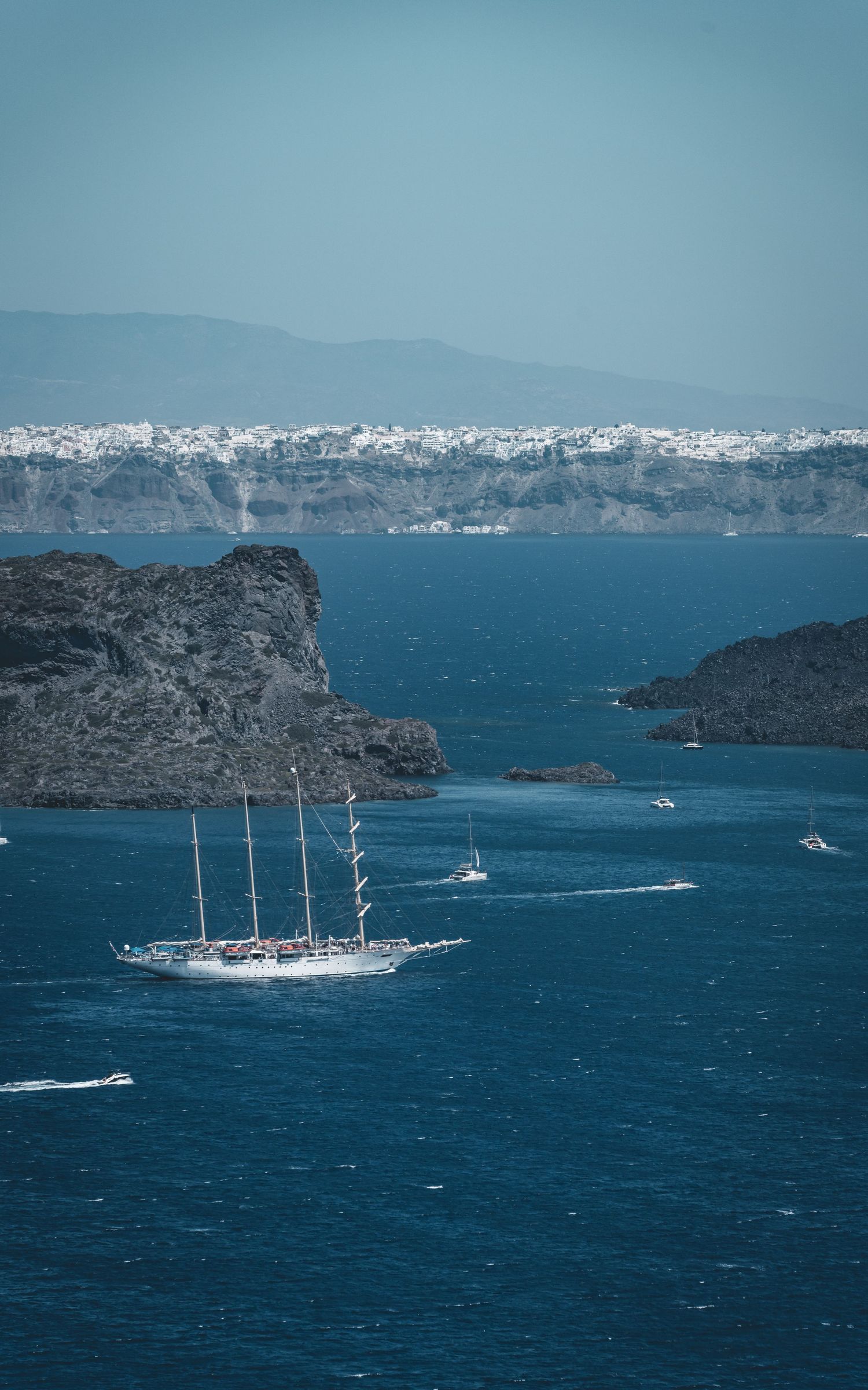 Tall sailing ship navigating blue waters near volcanic rocky islets with white Santorini village beyond.