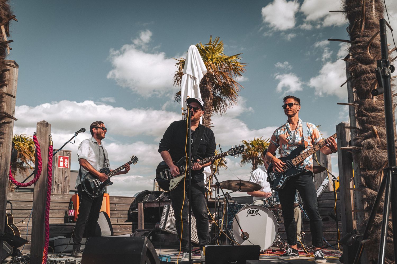 Live band performing outdoors on a beach stage with palm trees and cloudy sky backdrop.