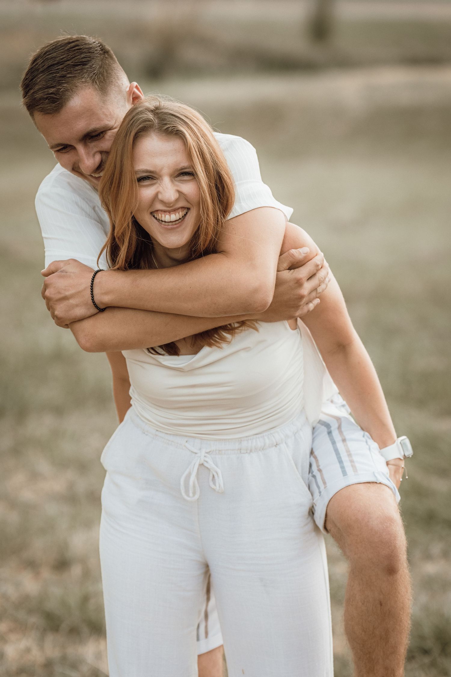 Happy couple embracing outdoors, man hugging woman from behind as she laughs joyfully in a sunny field.