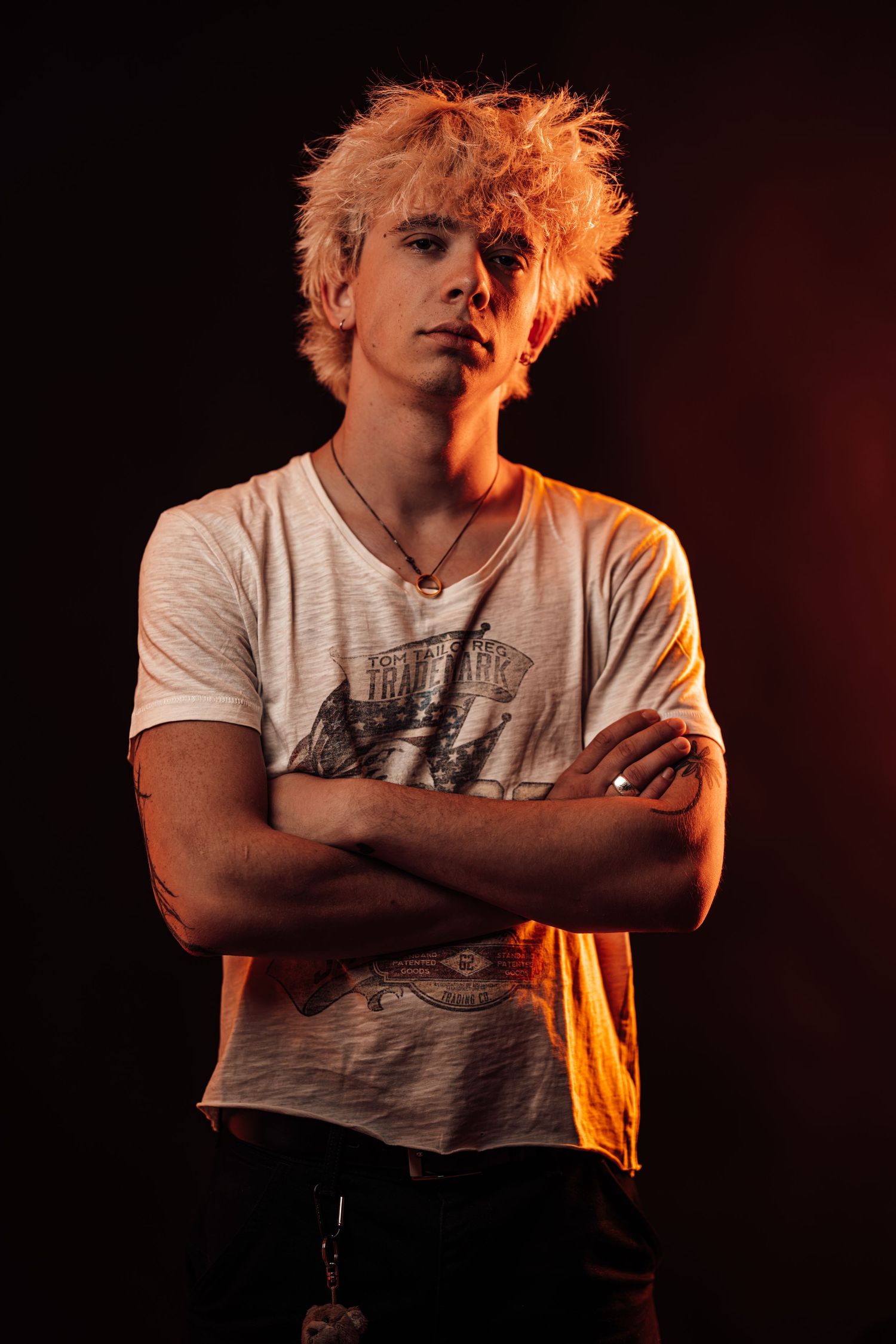 Young man with bleached hair, arms crossed, wearing a graphic tee, lit by warm orange light against dark background.