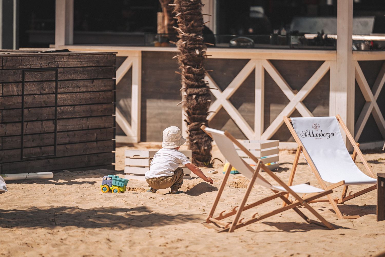 Toddler playing in sand beside deck chairs outside a beachside restaurant.