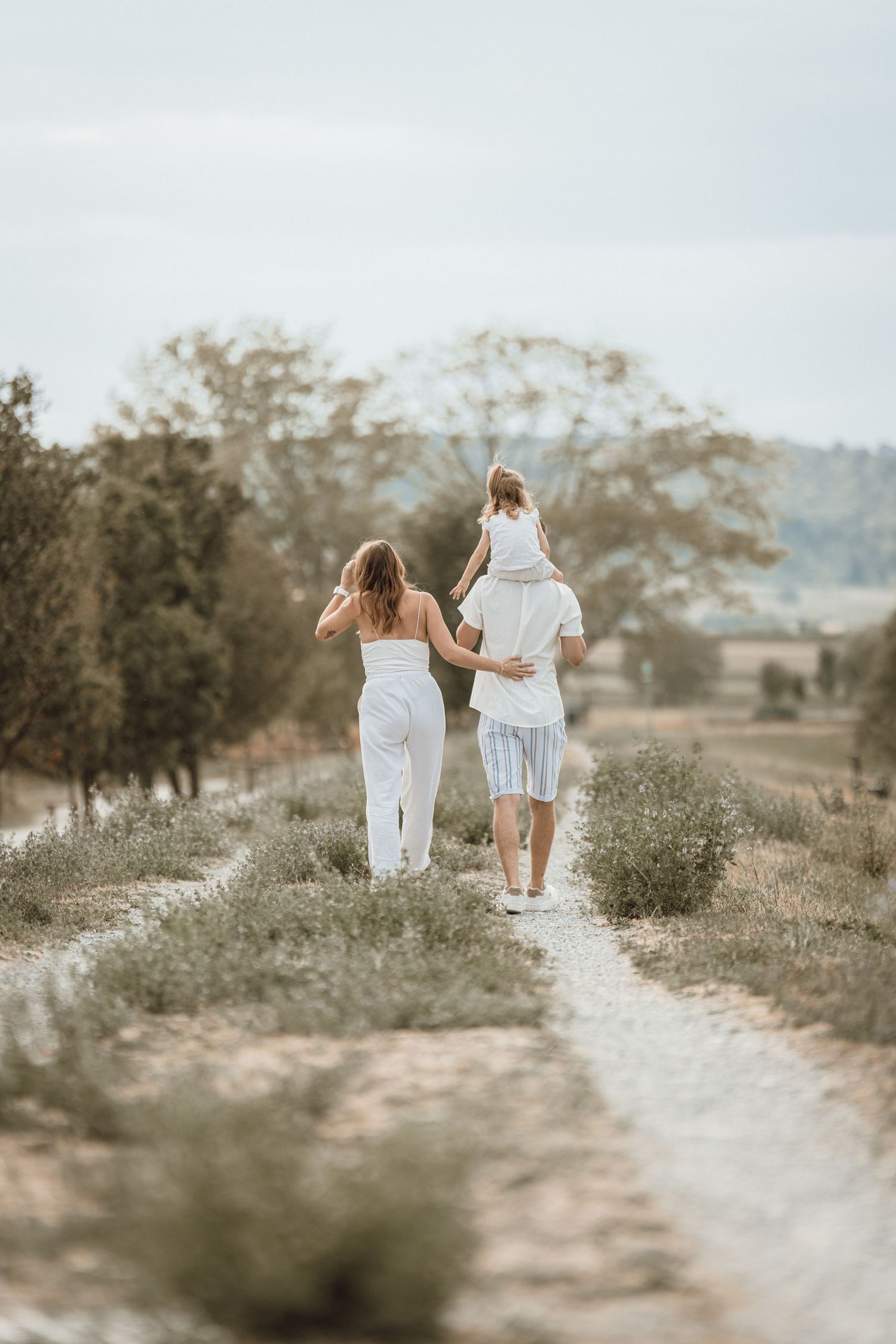 Family in white outfits walking down a rustic countryside path, with child sitting on parent's shoulders.