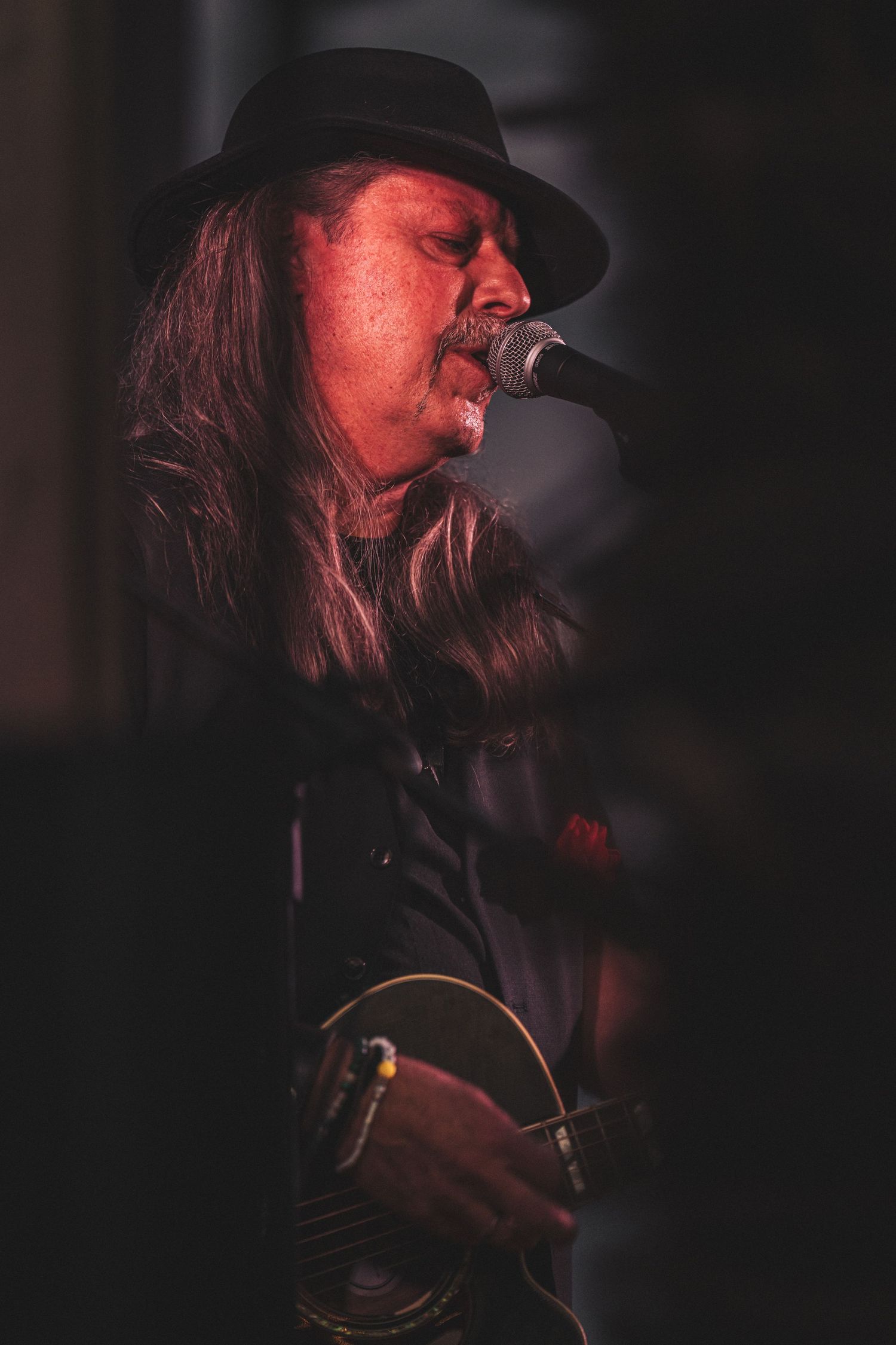 Long-haired male singer in black hat performing at microphone with guitar in red stage lighting.