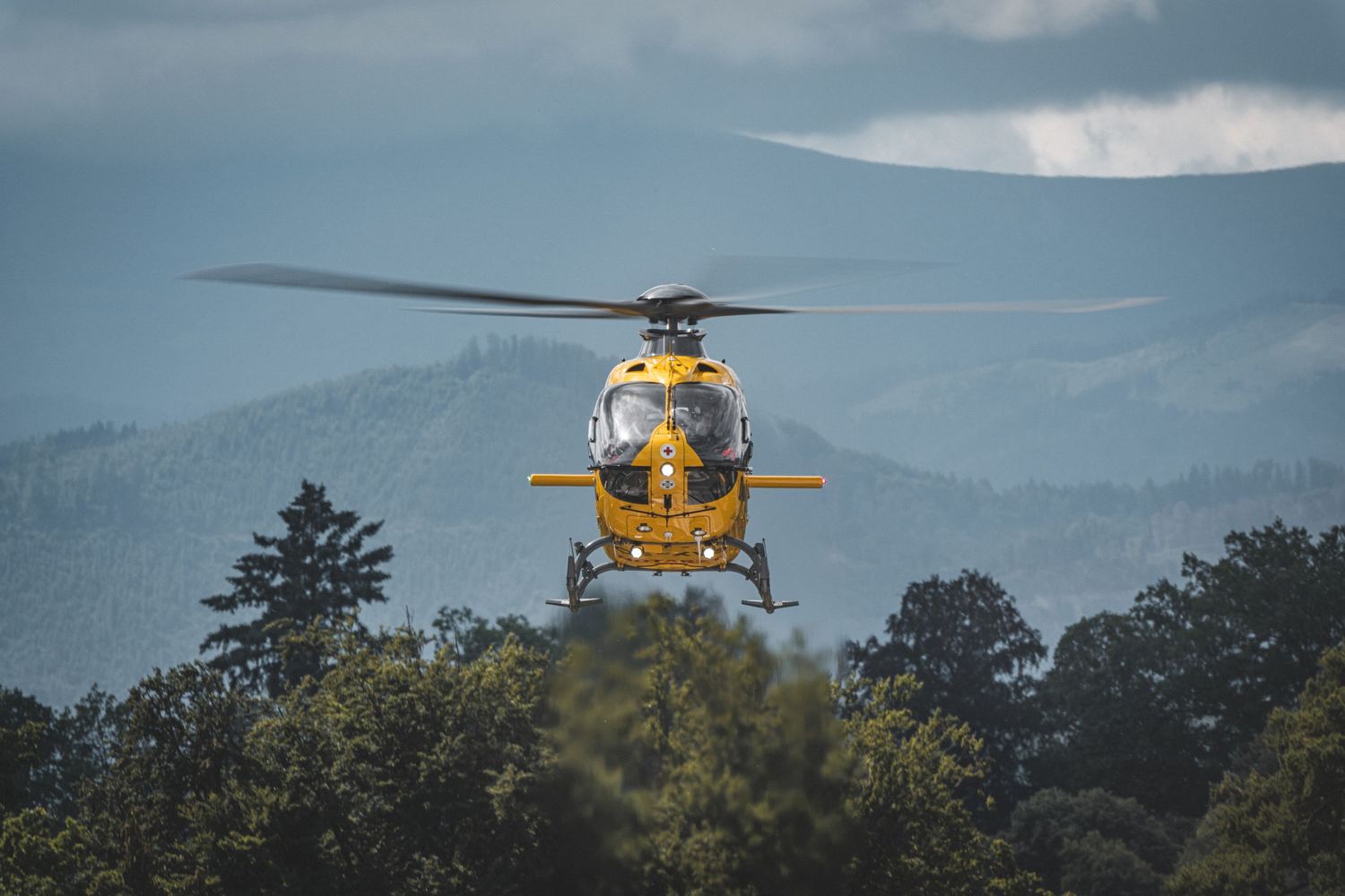 Yellow rescue helicopter flying low over treetops with misty mountains in the background.